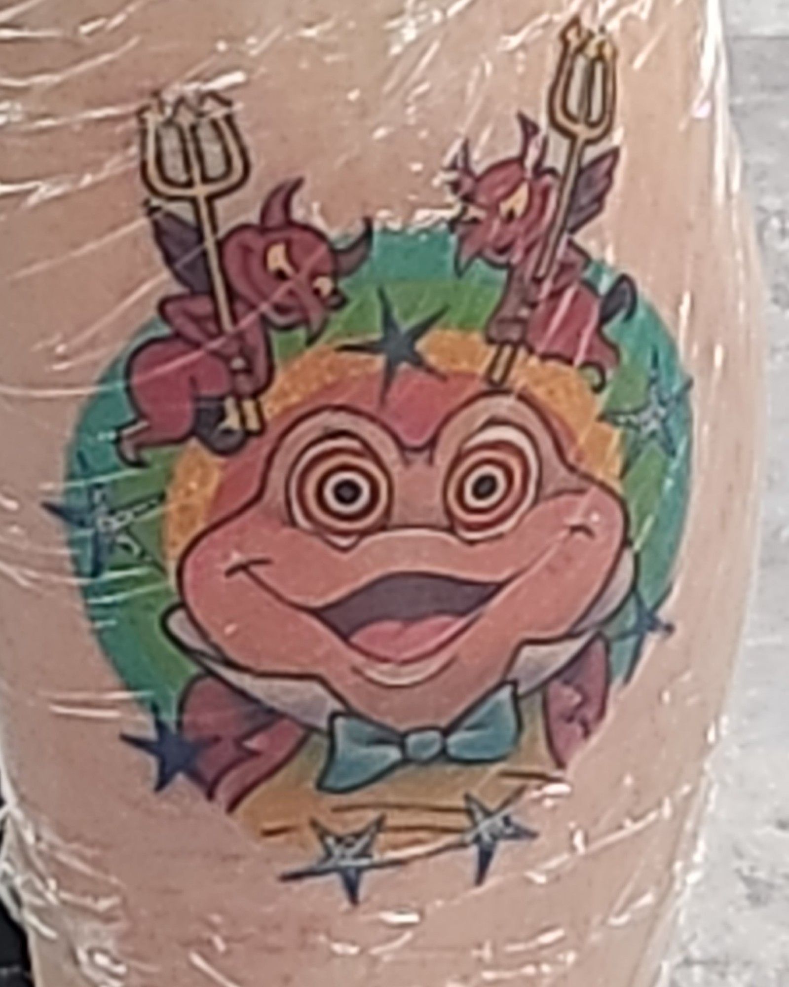 A plastic covered tattoo of Mr Toad with swirly eyes and two devils egging him on