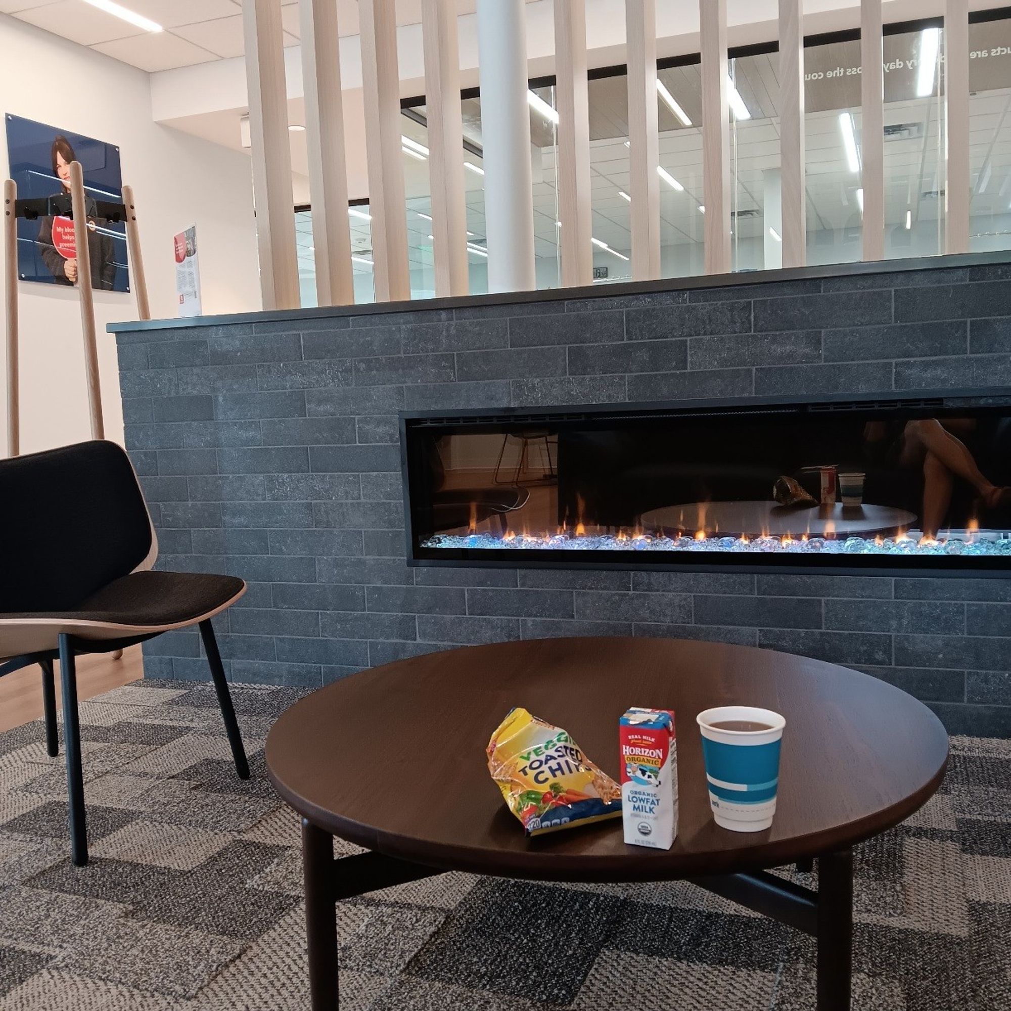 Brightly-lit lounge with snacks and drinks, chair and table in front of a decorative fireplace. Blood donation posters on the wall.