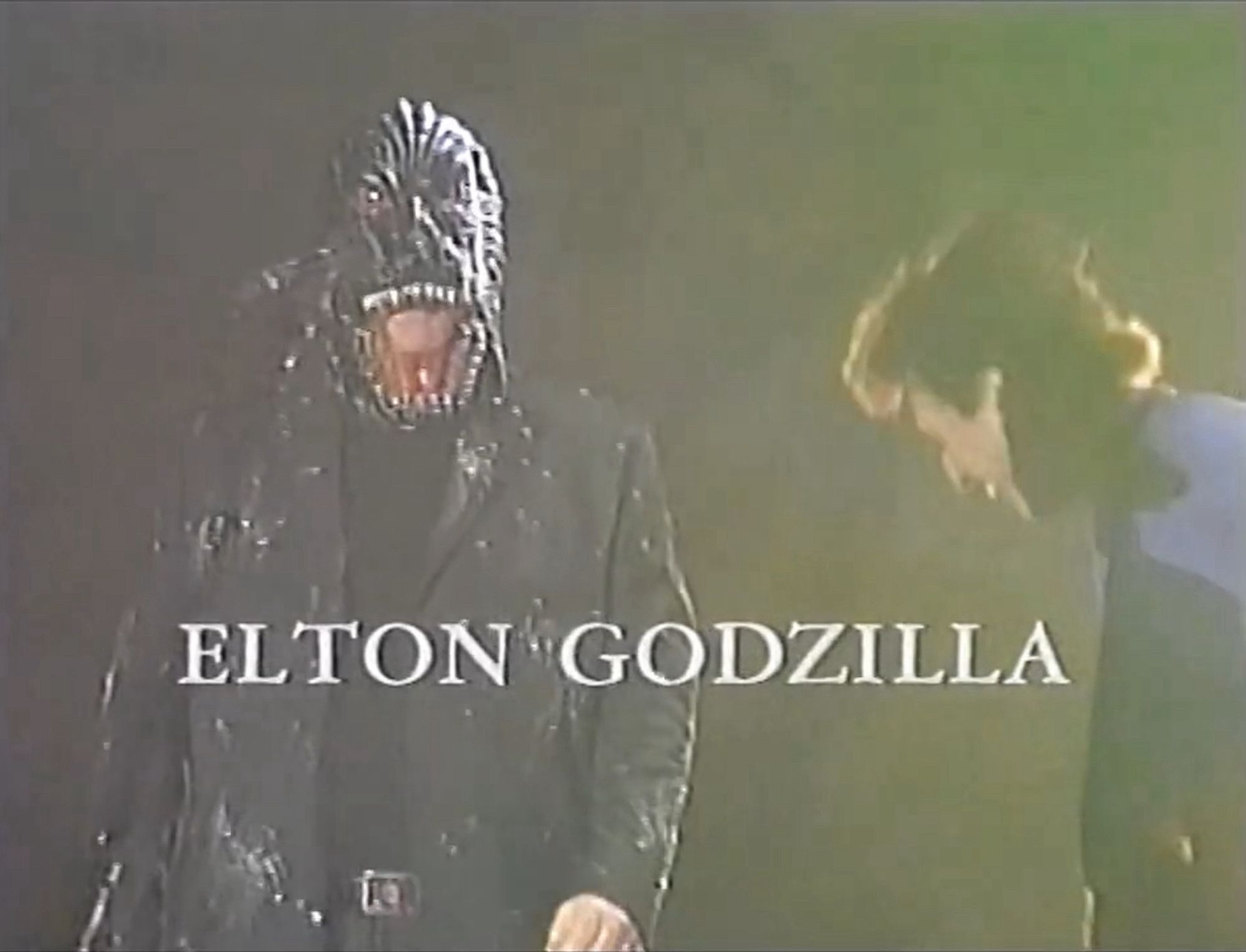 Elton John wearing a Godzilla mask walking onto stage in Tokyo with the words “ELTON GODZILLA” superimposed.