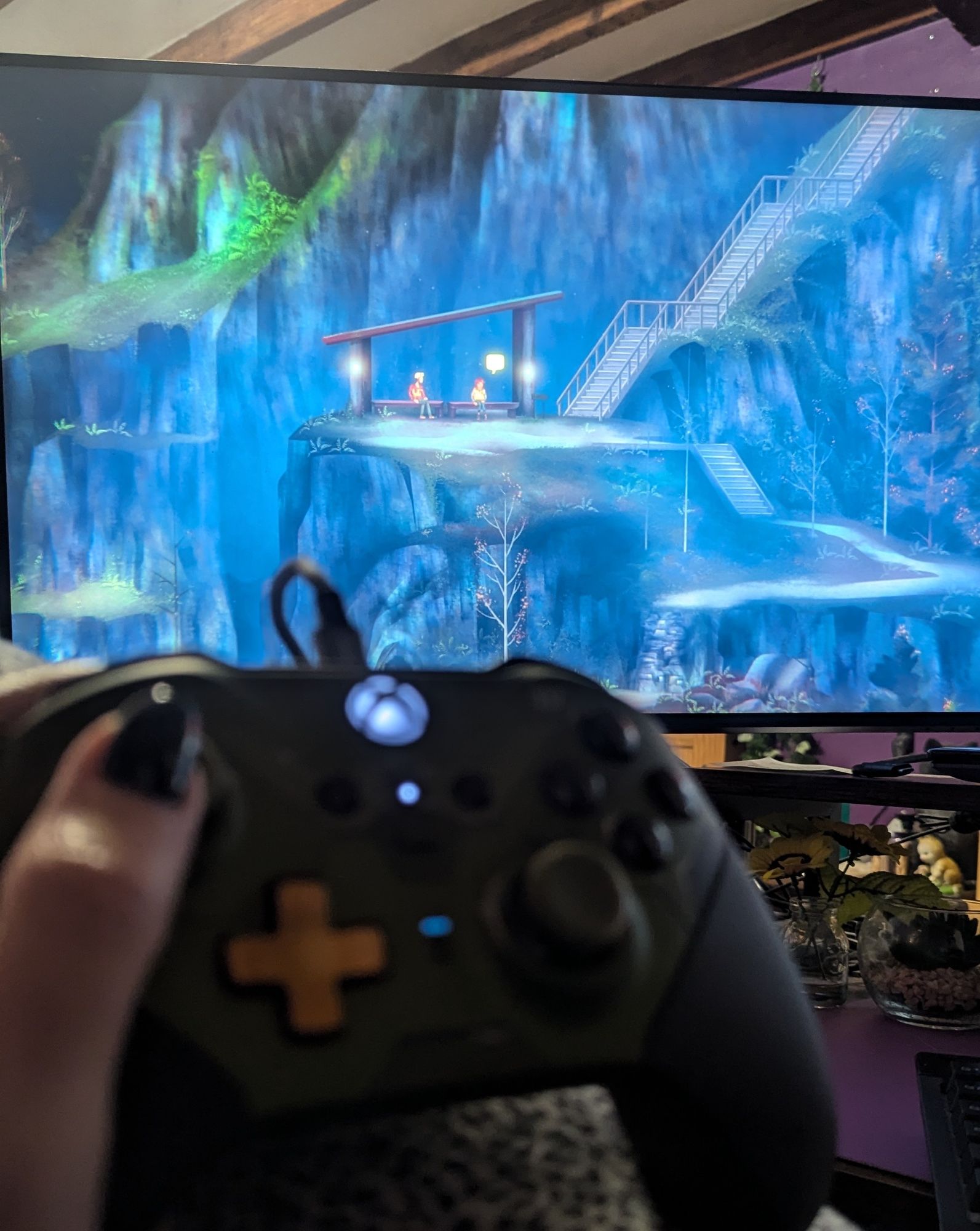 Jo's left hand holds and Xbox controller infront of a large PC screen. On the screen is a stylised clifftop forest ath with two small figures sat in a wooden bench.