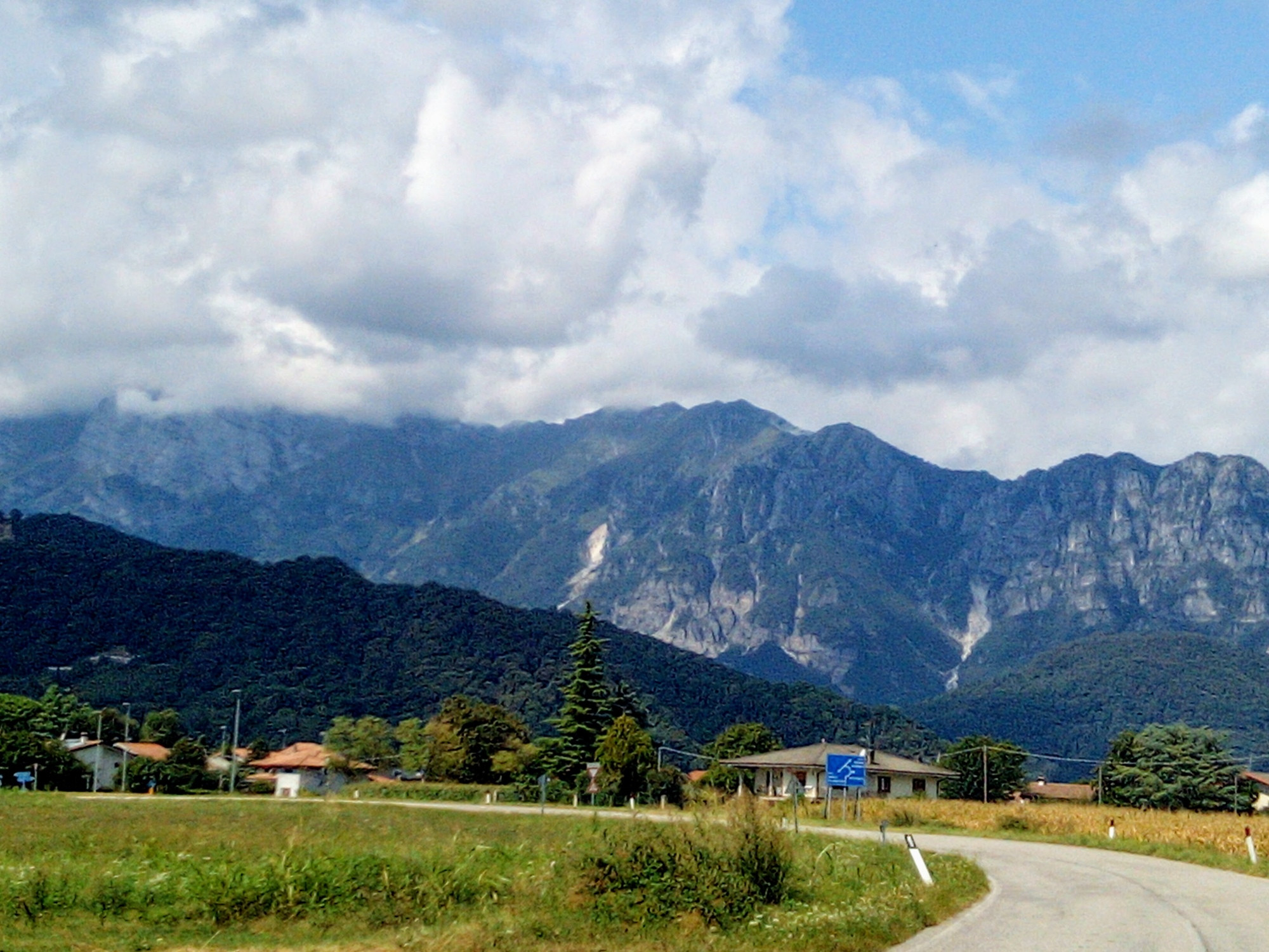 A view of mountains under a cloudy sky. A grey road curves from the right hand corner to the left. Houses in the mid ground among trees. In the left foreground a green field. 