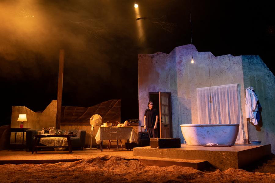 The set of Desert Stories for Lost Girls, the fractured interior of a modest home, with dirt filling the stage below the platform, and an image of the sea projected across the fractured wall. 