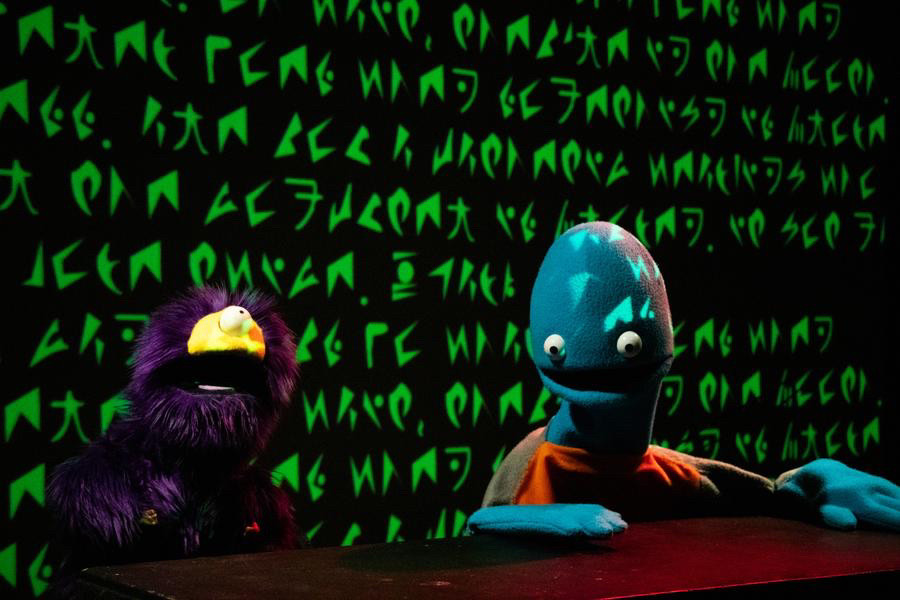 Two alien puppets from Happy Puppet Hour 2, seated behind a desk, shocked expressions on their face as an error message in a bright green alien font is projected across the wall behind them.