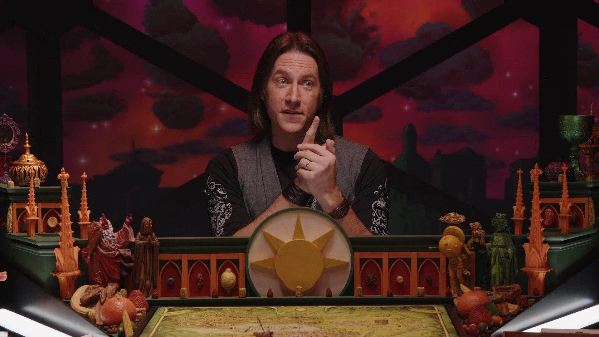 Matthew Mercer on the set of Dimension 20's Ravening War, behind the DM screen of spires and arches with assorted fruit imagery, and with the projected dusk skyline under fruit-shaped clouds on the dome behind him.