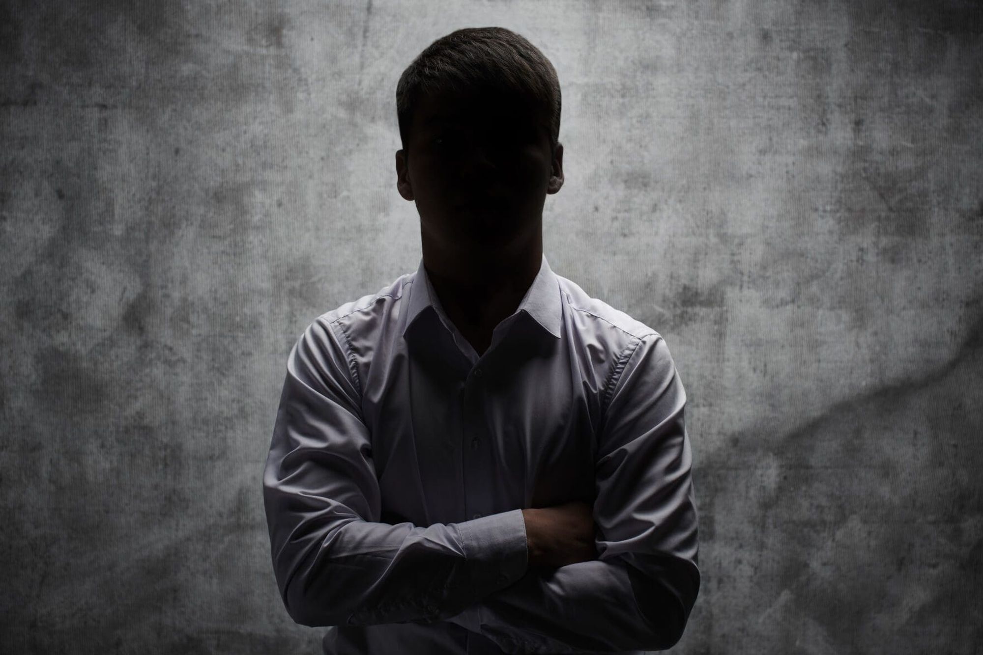 A youngish man standing erect arms folded over his chest wearing a light-colored format shirt. He is lit from behind with his head in particular obscured by darkness, in front of a mottled gray wall. In other words, a sterotypical anonymous source picture.