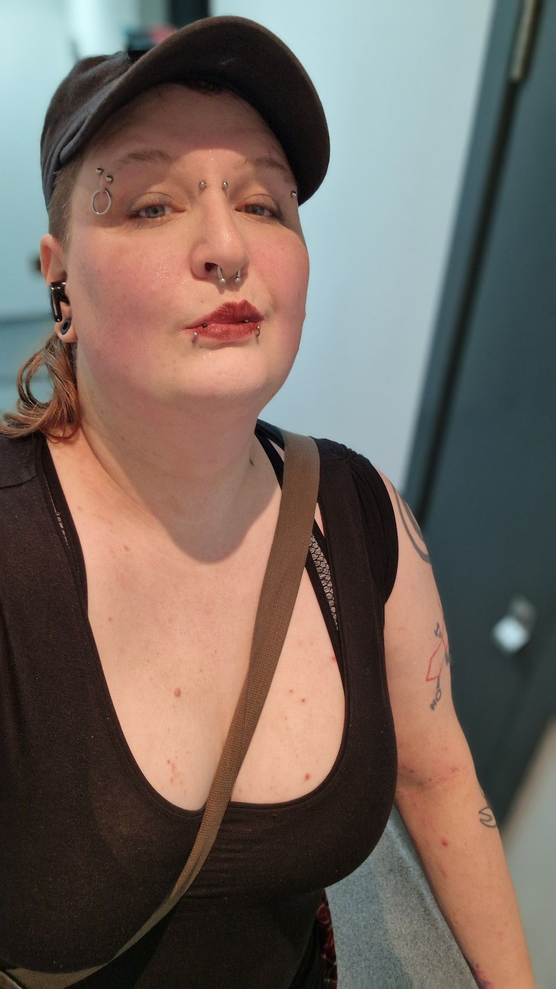 Krissy, a white trans woman with piercings. She's wearing a black scoop-neck top and a black baseball cap.