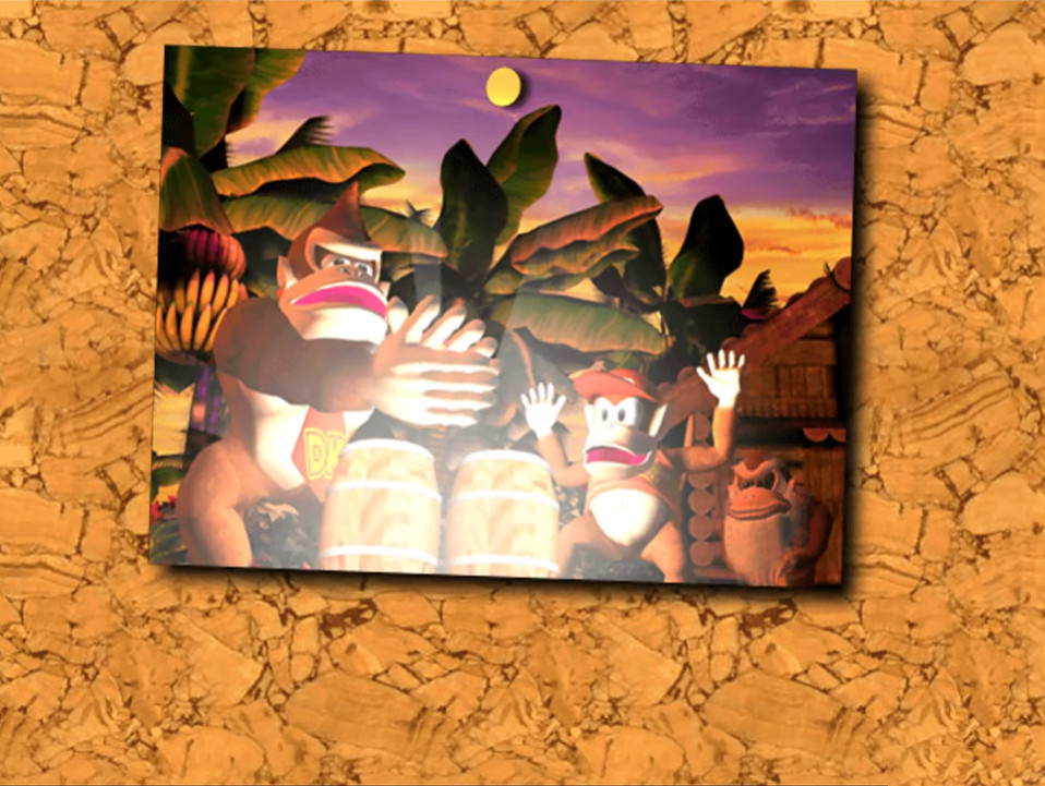 A photograph on a corkboard of Donkey Konga PAN PANNING (clapping) above the DK Bongos outside of Cranky's Cabin, as Diddy and Cranky look on as the bongos begin to glow. Screenshot from Donkey Konga.