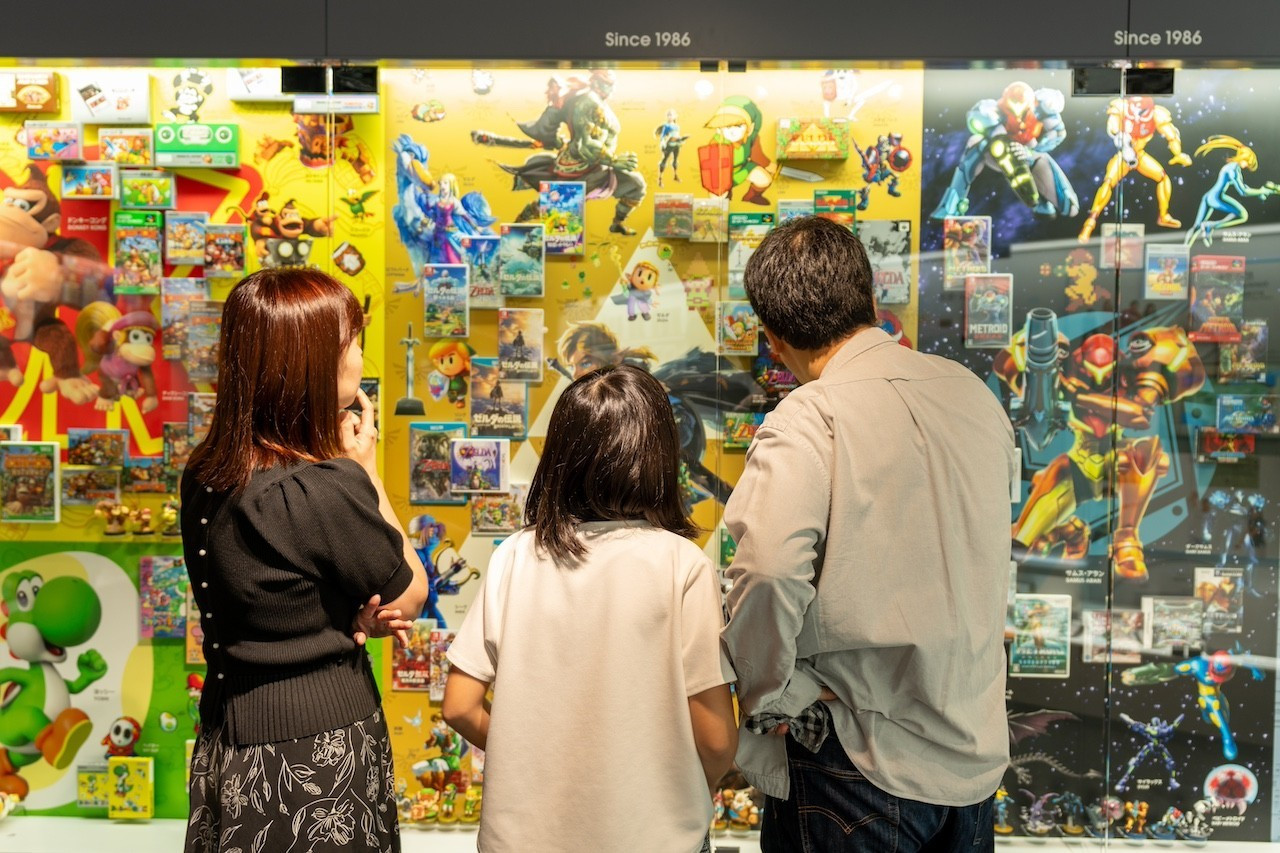 Three guests look at Nintendo series timelines in a display case at the Nintendo Museum in Kyoto. Includes the Donkey Kong series, Yoshi series, Legend of Zelda series, and Metroid series.