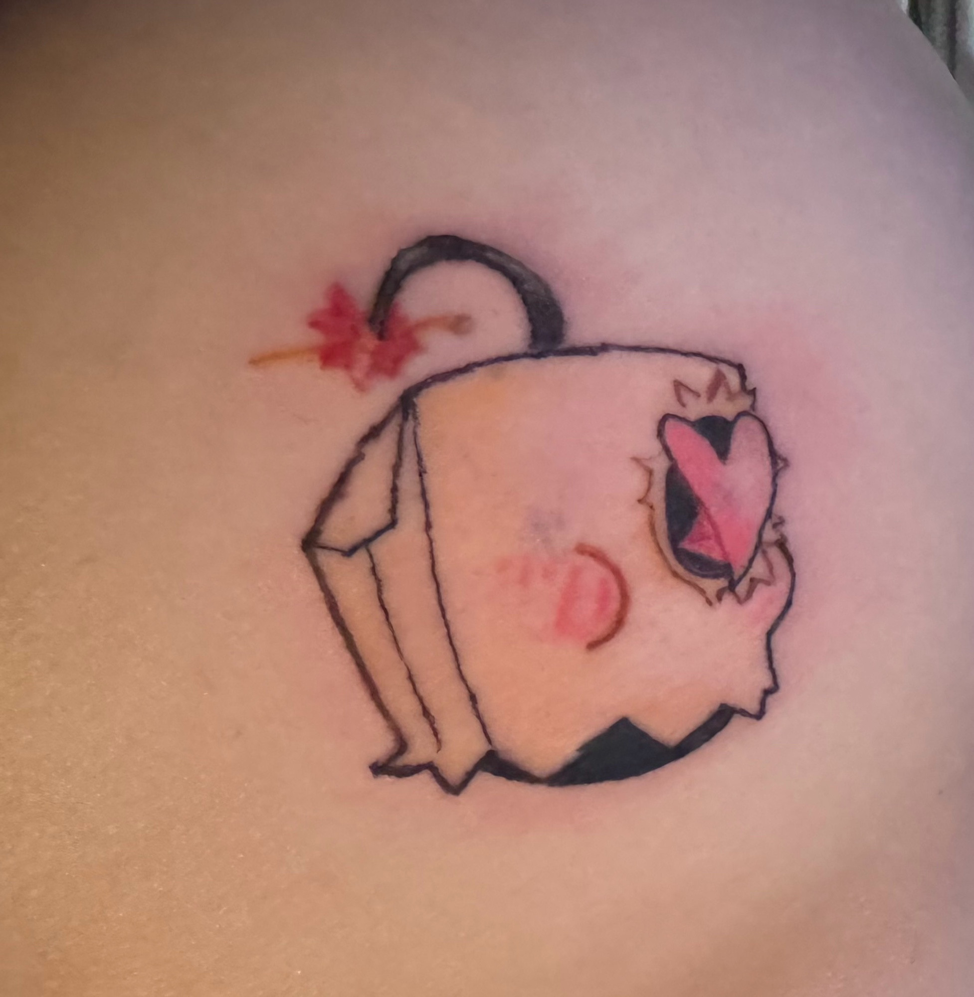 A tattoo of the fighting game move called “take my love” that looks like a brown paper bag with a fuse on top and an eye hole cut out with a heart coming out of the hole. The bag also has rosy cheeks. The entire image is the head of Doctor Faust, a fighting game character in guilty gear