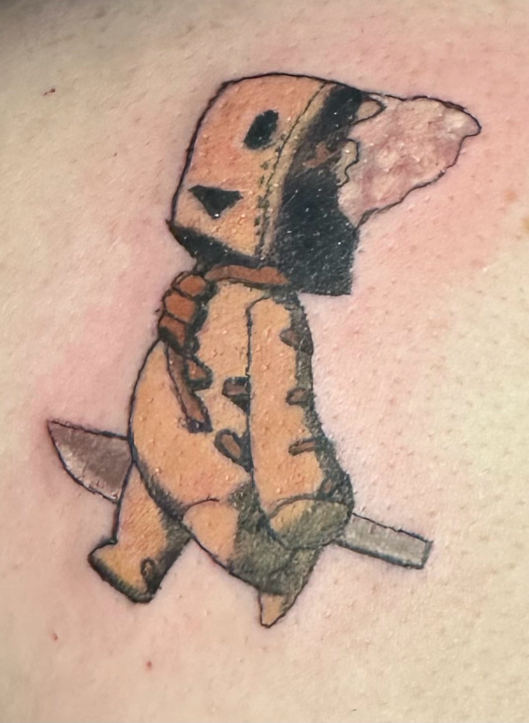 Image of a tattoo of Doctor Faust’s familiar Mini Faust. Mini Faust is a rounded arm and legged doll that is made from a brown sack, he has a square head with one eye and a mouth hole. There is white stuffing coming out of the left side of his head, he has an orange noose like necklace and orange cross stitching up both outer arms and both sides. He is holding a small silver scalpel and walking to the left 