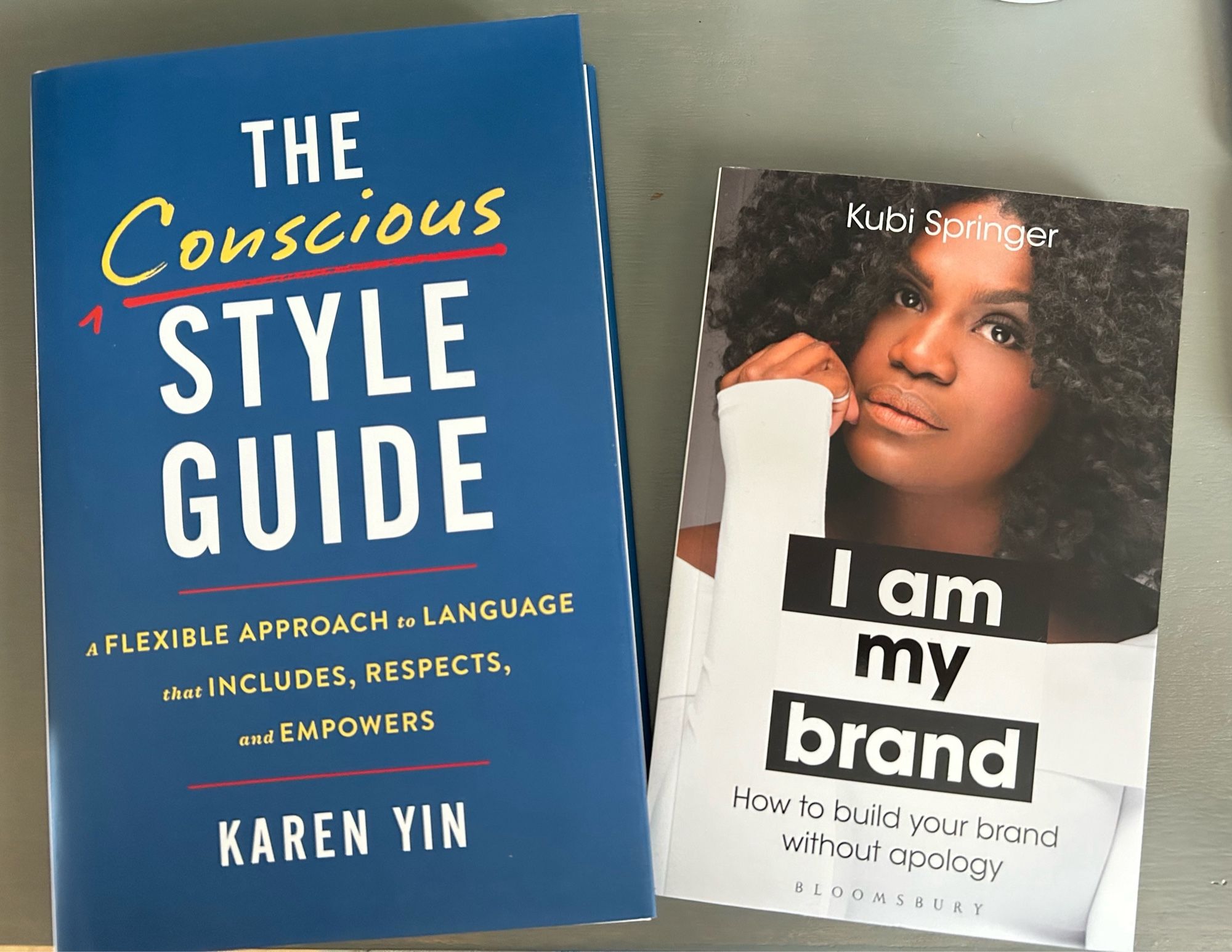 Two books sit on a grey desktop: The Conscious Style Guide: A Flexible Approach to Language that Includes, Respects, and Empowers by Karen Yin, and I Am My Brand: How to Build Your Brand Without Apology by Kubi Springer.