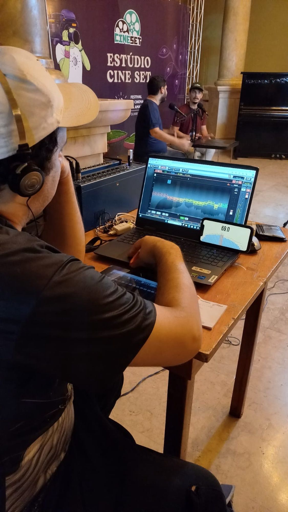 Um homem sentado de costas usando um computador controla uma mesa de som, e à frente acontece uma entrevista no hall do Teatro Amazonas