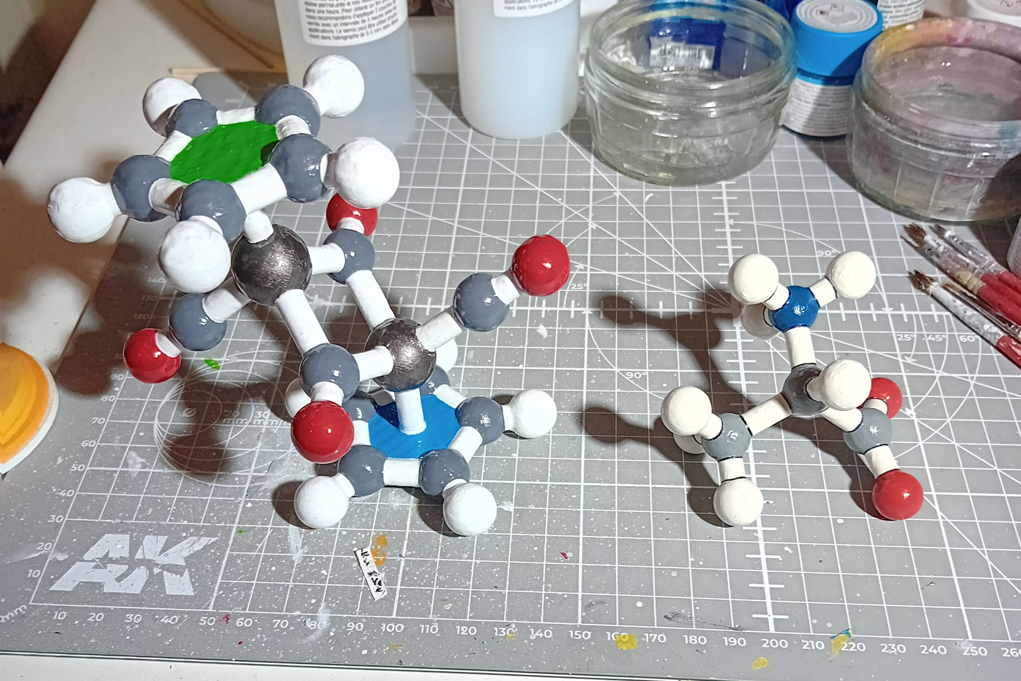 Picture of two hand painted 3d printed crystal structures, a glossy [(Cp)Fe(CO)2]2 with coloured infill on the rings, and an alanine zwitterion painted in matt style with black highlights between the atoms and bonds