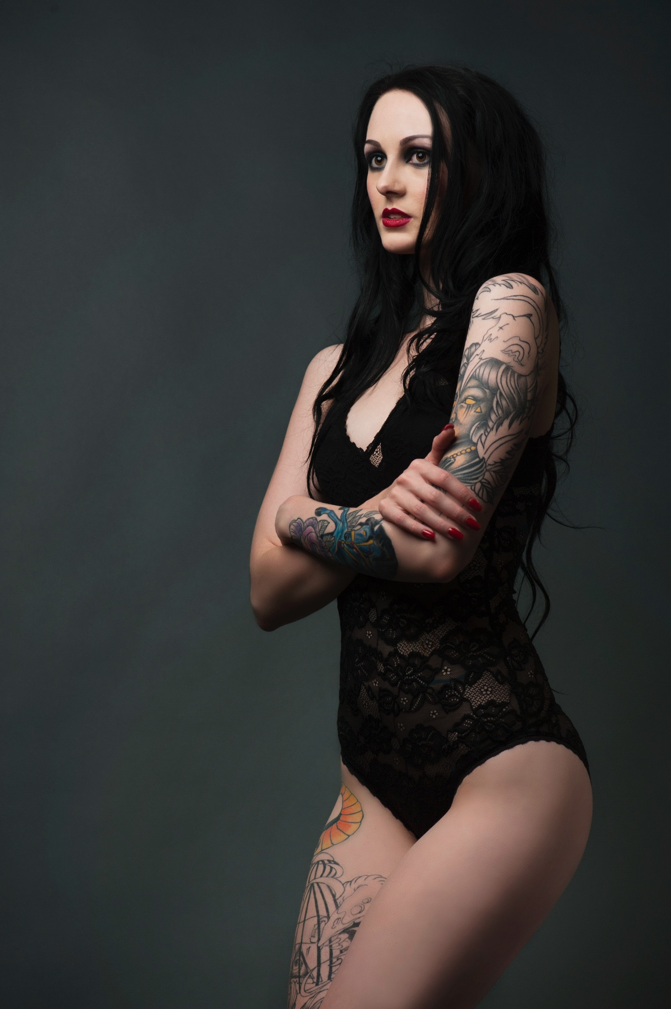 A woman with long, wavy black hair stands confidently against a dark, moody background. She is wearing a fitted black lace bodysuit that accentuates her figure, while her arms are crossed over her chest, giving a composed yet fierce expression. Her makeup adds to the gothic vibe, with dark eyeliner framing her eyes and bold red lipstick providing a striking contrast. Her tattoos are prominent, covering her right arm and extending to her hip, featuring intricate designs with bold colors, including a dragon and various other elements. Her nails are painted red, complementing her makeup and adding to the powerful yet elegant look. The overall mood of the image is one of empowerment and bold self-expression, mixing alternative fashion with a classic, sophisticated style