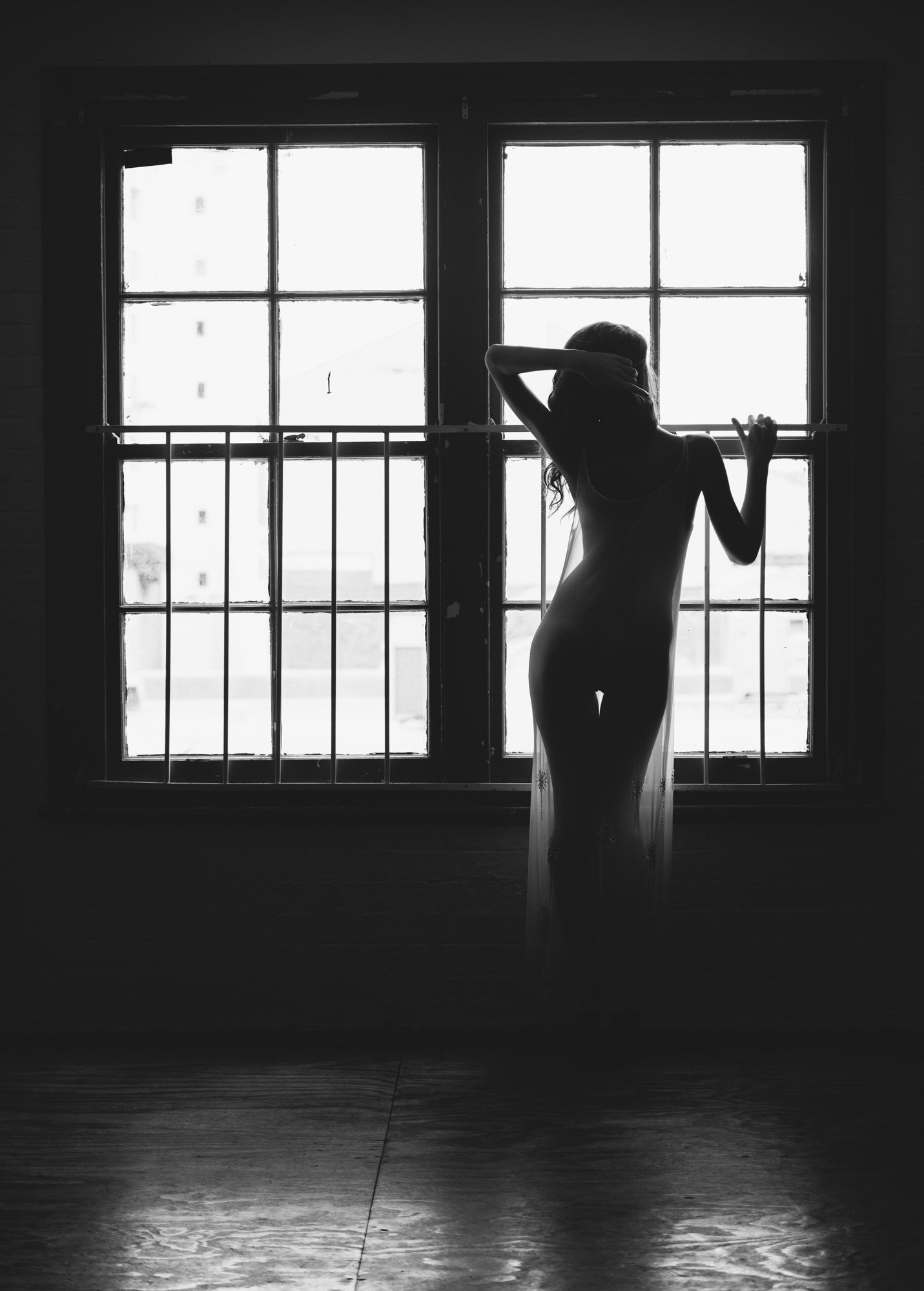 A black-and-white photograph shows a woman standing in front of a large window, her body in silhouette. She is dressed in a sheer, flowing fabric, her form subtly revealed by the soft light streaming through the window behind her. One arm is raised, resting on her head, while the other gently grasps a window bar. The window has metal bars running horizontally across the panes. The room is dimly lit, emphasizing the contrast between the bright light from outside and the shadows within. The image evokes a quiet, contemplative mood, with the interplay of light and shadow creating a sense of stillness and introspection. The woman’s posture and the simplicity of the setting enhance the emotional tone of solitude and calm. The overall atmosphere is peaceful, yet there’s an air of mystery, as the viewer is drawn into the tranquil moment.