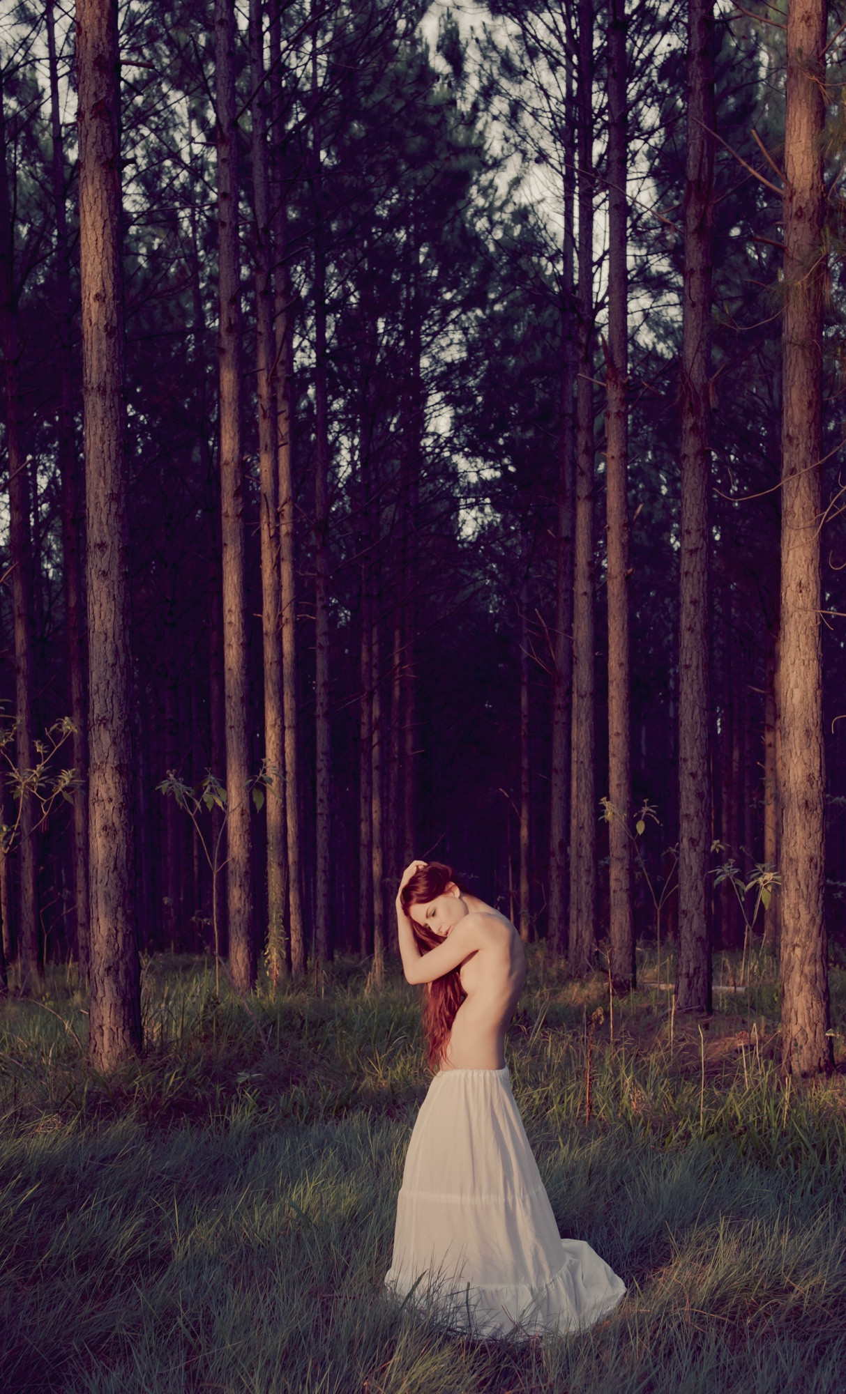 A serene outdoor scene featuring a nude woman standing in a forest of tall pine trees. She is facing slightly downward, with one hand resting on her head and the other hand by her side. Her long, loose hair cascades over her shoulder, partially covering her body. She is wearing a long, white flowing skirt that reaches the grass below. The soft, natural light filters through the trees, creating a peaceful and intimate atmosphere. The muted tones of the forest contrast with the warmth and vulnerability of the human form.