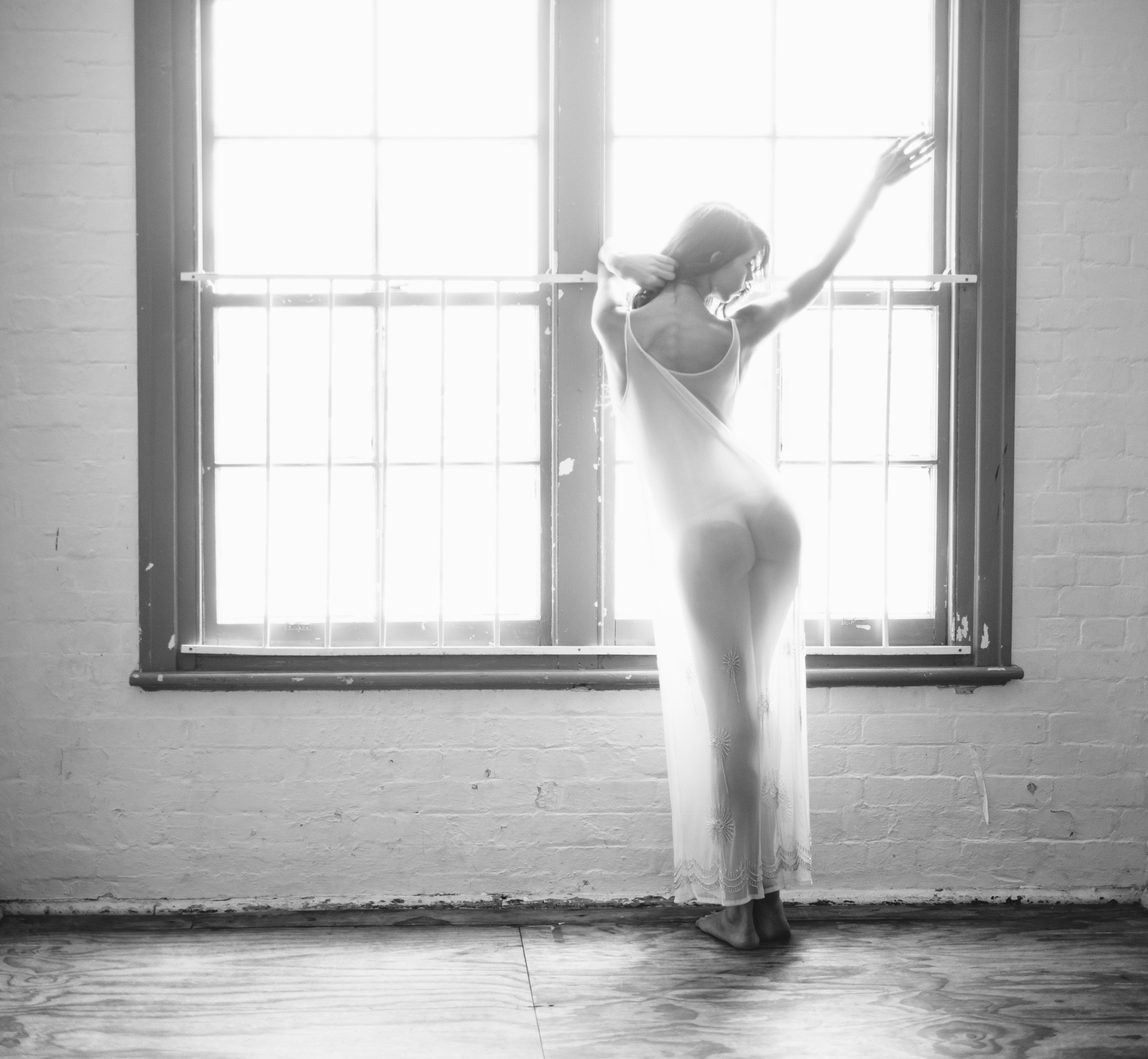 A black-and-white photo of a woman standing barefoot in front of a large window with sunlight streaming in. She faces away from the camera, her body draped in a sheer, flowing fabric that subtly reveals her form. One arm reaches upward, while the other gently touches her hair. The bright light from the window creates a soft, ethereal glow around her silhouette, emphasizing the curves of her body and the texture of the fabric.