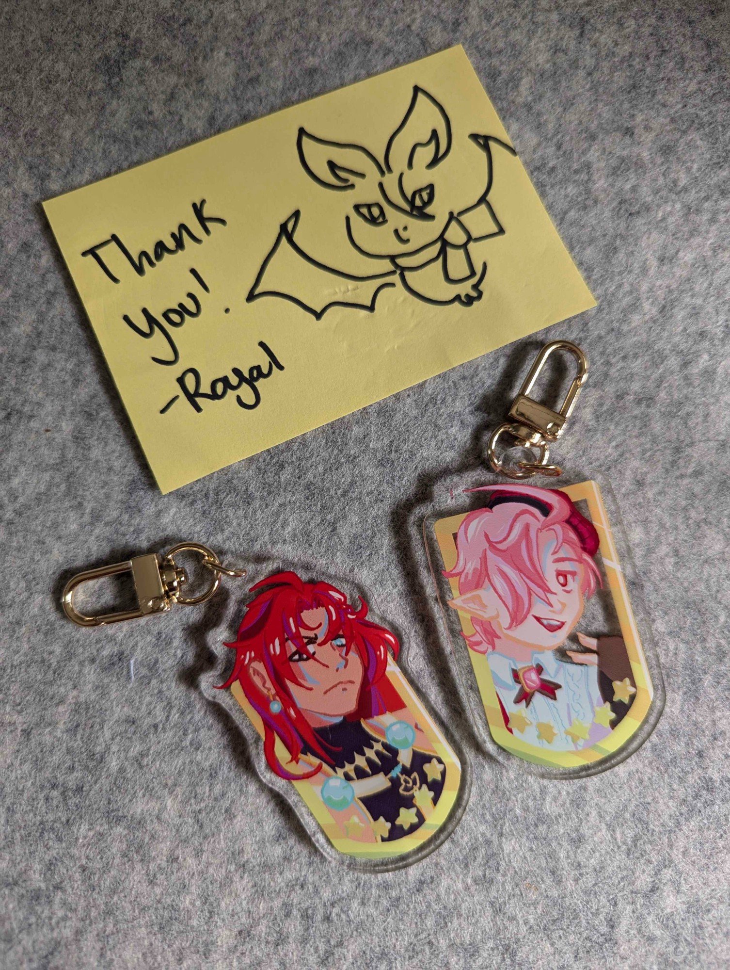 keychains featuring art of Aster and Dante from nu: carnival. a yellow note says "Thank you! Royal" and has a sketch of Aster in his bat form