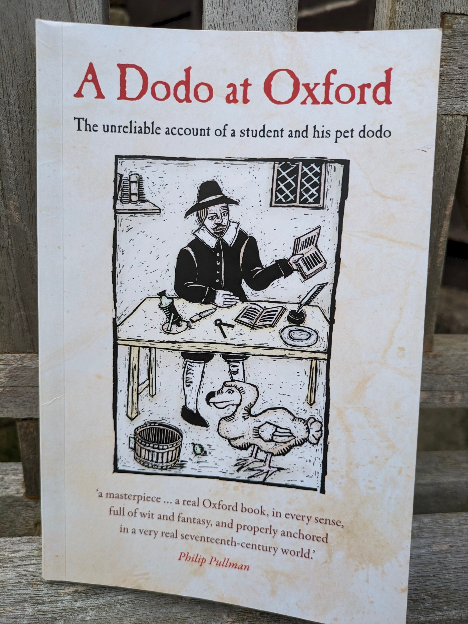 A book, called 'A Dodo at Oxford', subtitled 'The unreliable account of a student and his pet dodo'.  An image is on the cover in the style of a late 17th century print of a man standing behind a table costumed appropriately with a dodo in front of the table, bottom right of the image.

Underneath is a quote from Philip Pullman:  'a masterpiece a real Oxford book, in every sense, full of wit and fantasy, and properly anchored in a very real seventeenth-century world'