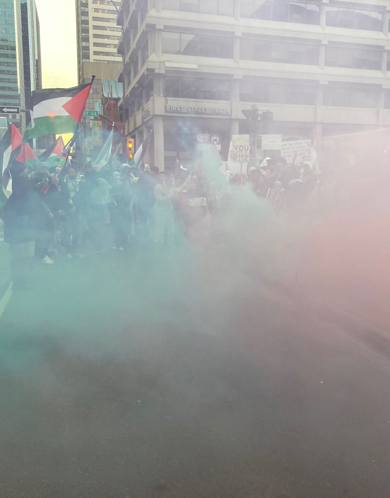 A picture of the Palestinian Solidarity Protest in Calgary on November 12th, 2023. Green- and red-coloured smoke is spreading across 4th Avenue. A number of Palestinian flags are visible on the other side of the smoke.