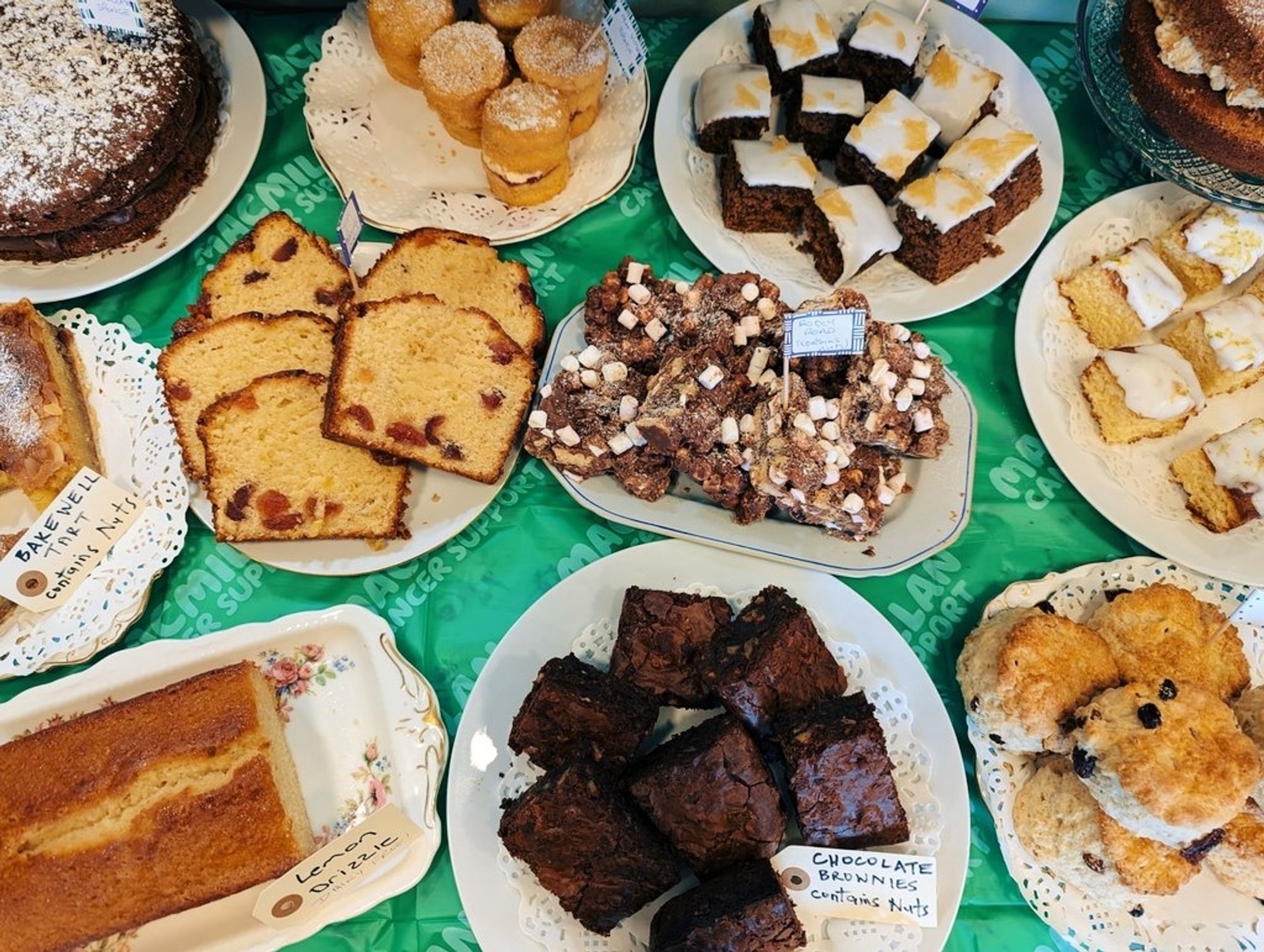 A selection of plated-up cakes, scones, brownies and tiffin for sale on a "Macmillan Cancer Support" table cloth