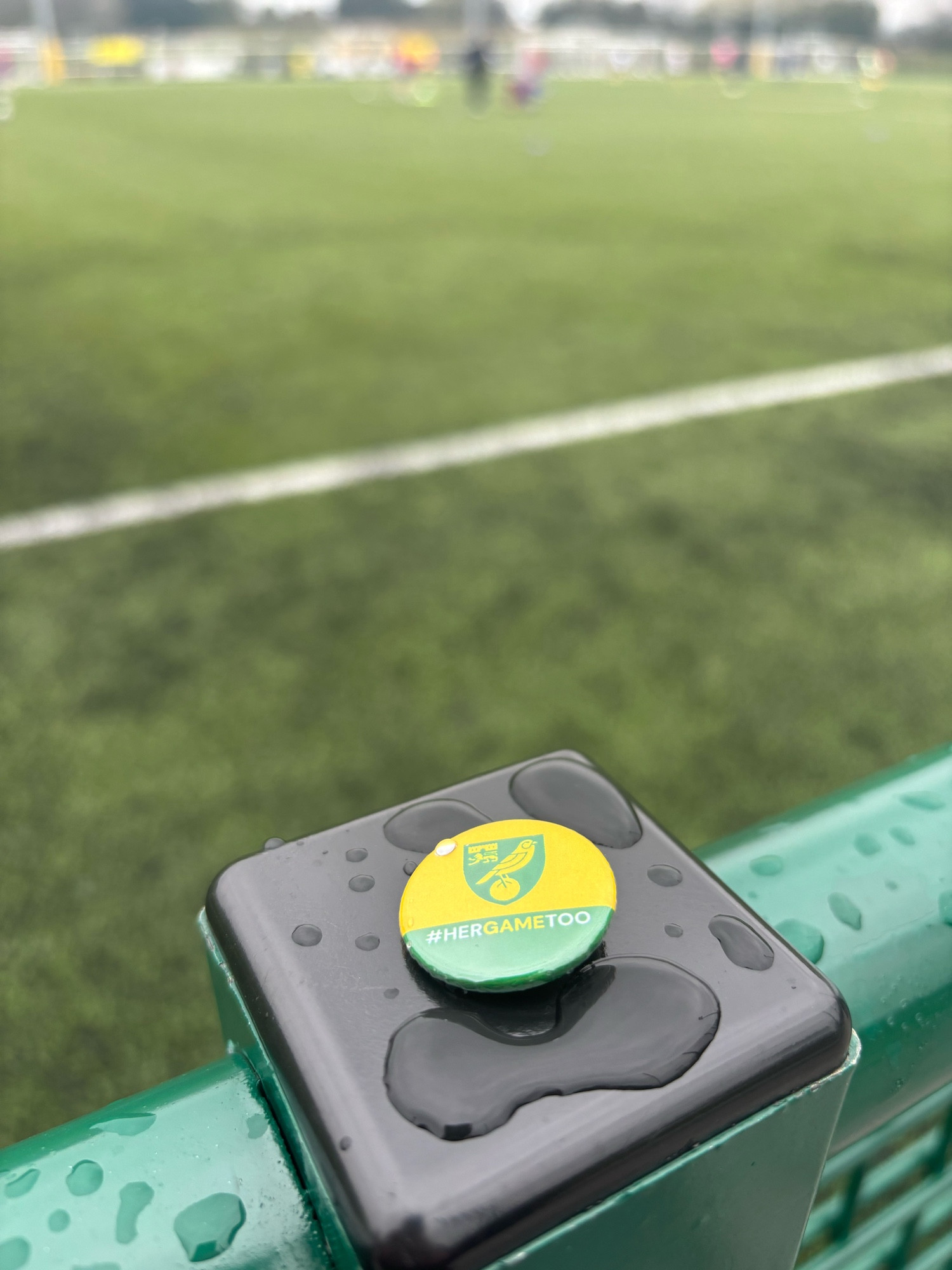 A #HerGameToo Norwich City button badge sitting on a wet fence post in front of the football pitch at The Nest ahead of todays Norwich City Women’s match