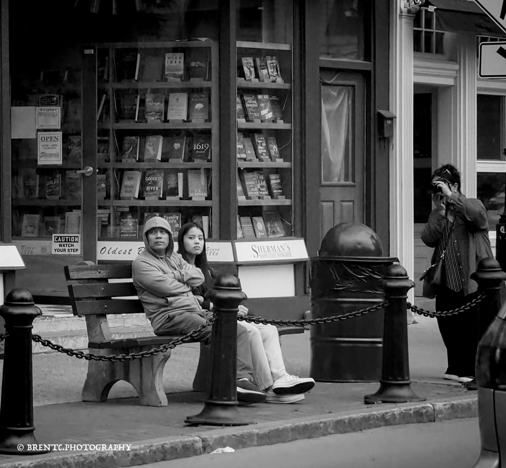 Black and white photo of two people sitting on a street bench in front of a bookstore and another photographer taking their photo at the same time.