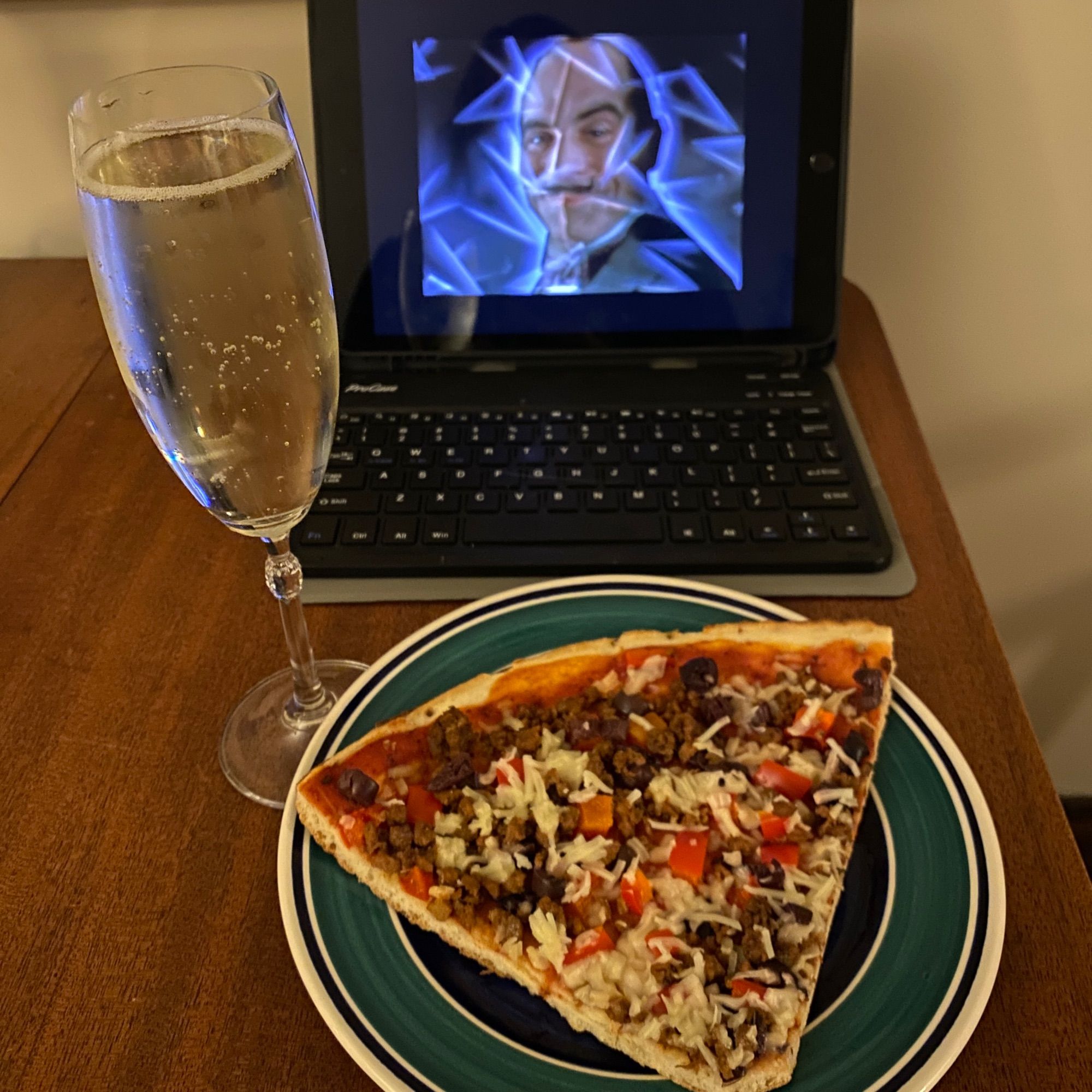 An iPad playing the opening credits of Agatha Christie’s Poirot starring David Suchet sits next to a small plate with a piece of pizza on it and a champagne flute full of Prosecco.