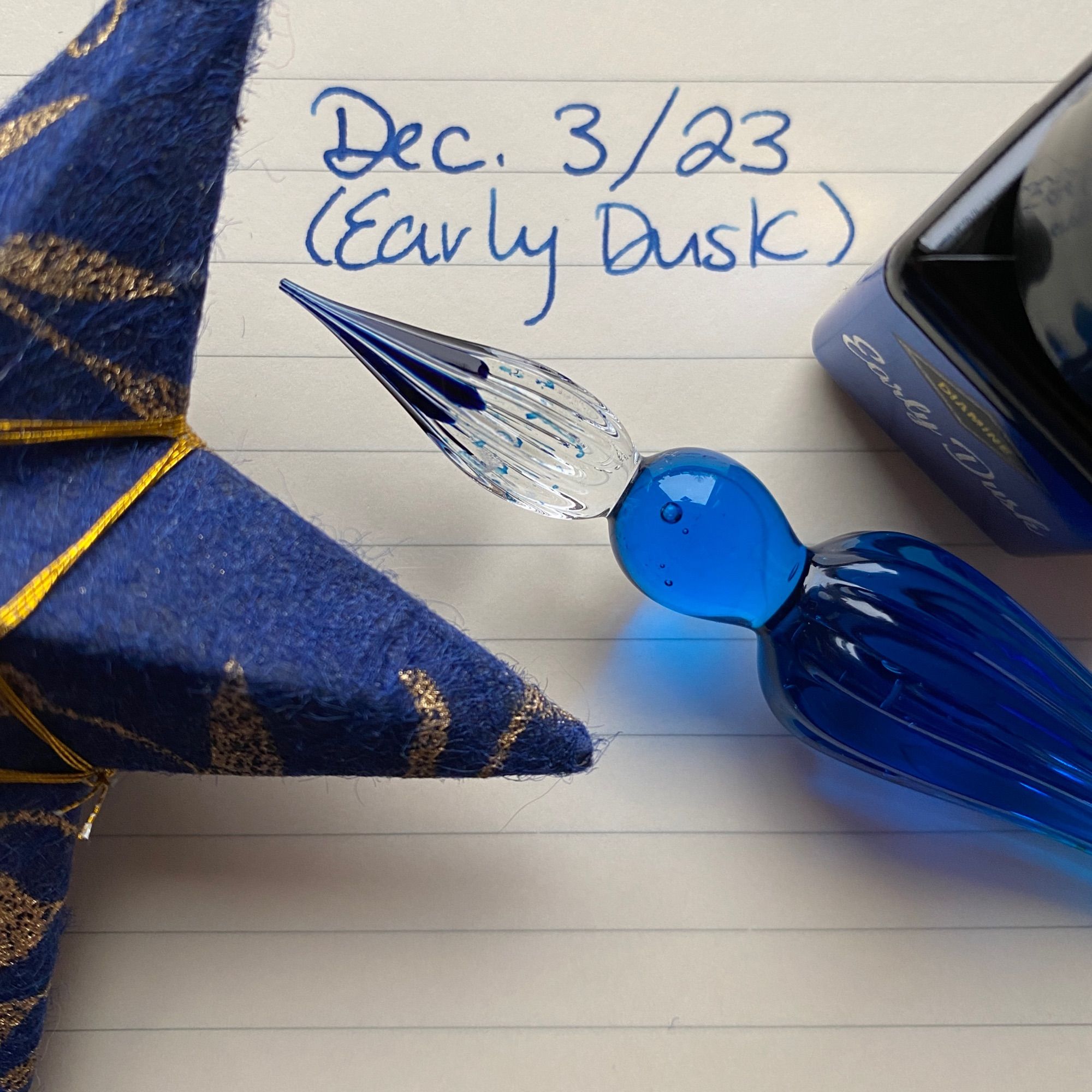 A lined page of a notebook with today’s date (December 3, 2023) written in blue ink. A blue glass dip pen, a small bottle of blue ink, and a blue and gold star ornament are on the page next to the words.