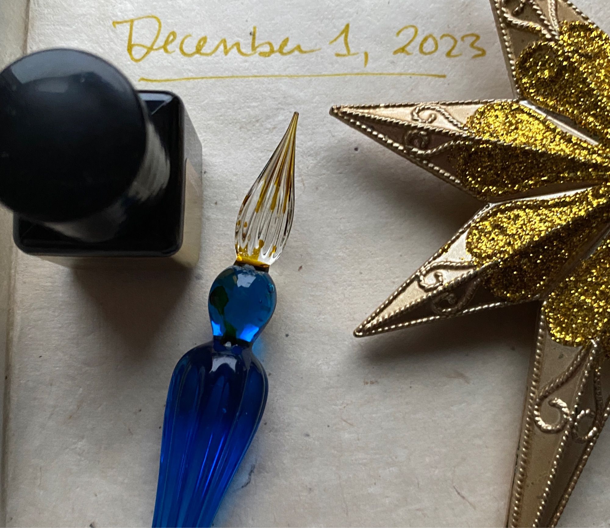 A notebook page with the date (December 1, 2023) written in gold ink. A small bottle of ink and a gold star holiday ornament sit on the page next to a blue glass dip pen.