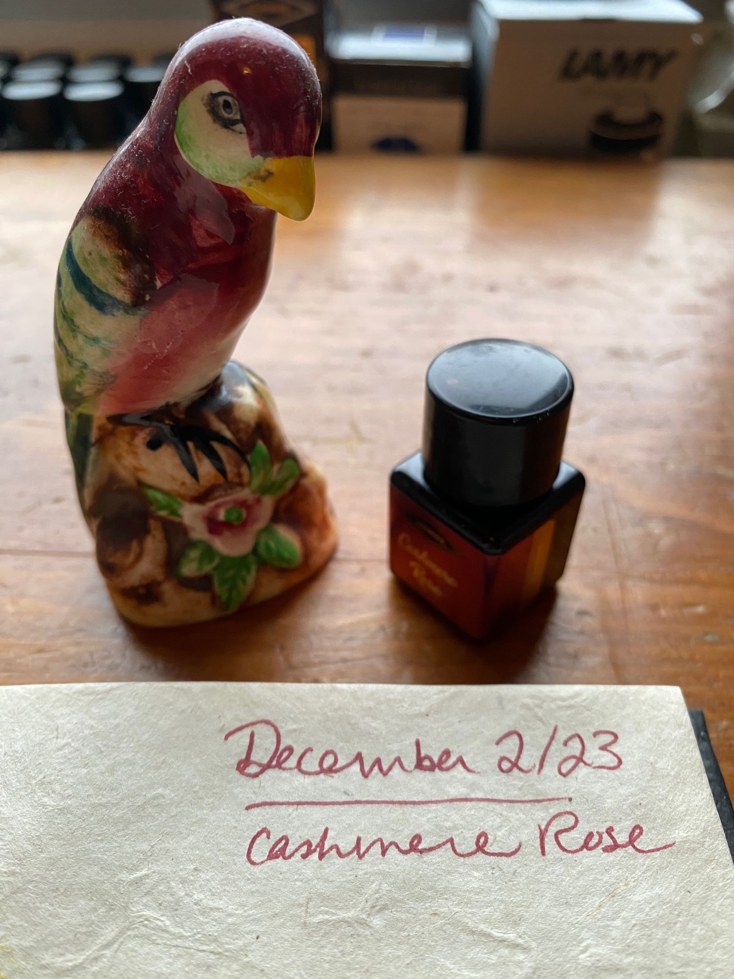 A notebook page with the date (December 2, 2023) and the words Cashmere Rose written at the top. A ceramic figurine of a red bird sits next to the notebook as does a small bottle of ink. The notebook is on a well worn wooden desk and in the background you can see other bottles of fountain pen ink.