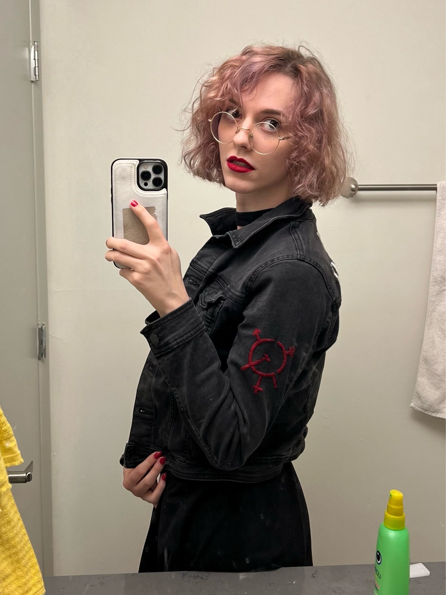 a mirror selfie of imy in a black denim jacket with the symbol of transgender marxism embroidered on the sleeve in red. her lipstick and nails are both red as well.