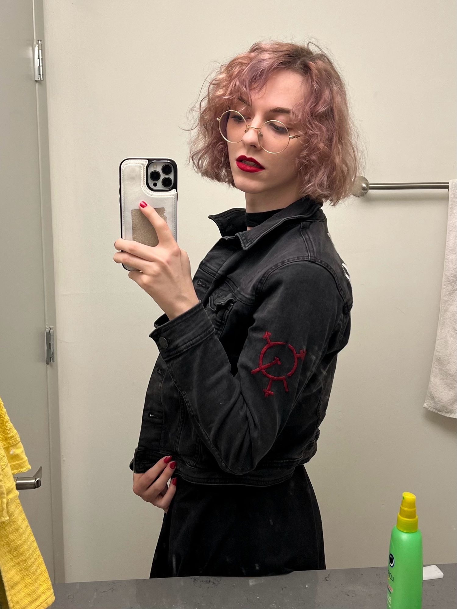 a mirror selfie of imy in a black denim jacket with the symbol of transgender marxism embroidered on the sleeve in red. her lipstick and nails are both red as well.