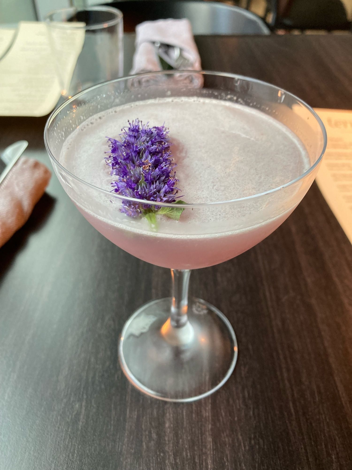 A purple-rose cocktail in a champagne coupe with purple flowers from mint I think floating on the frothy surface.