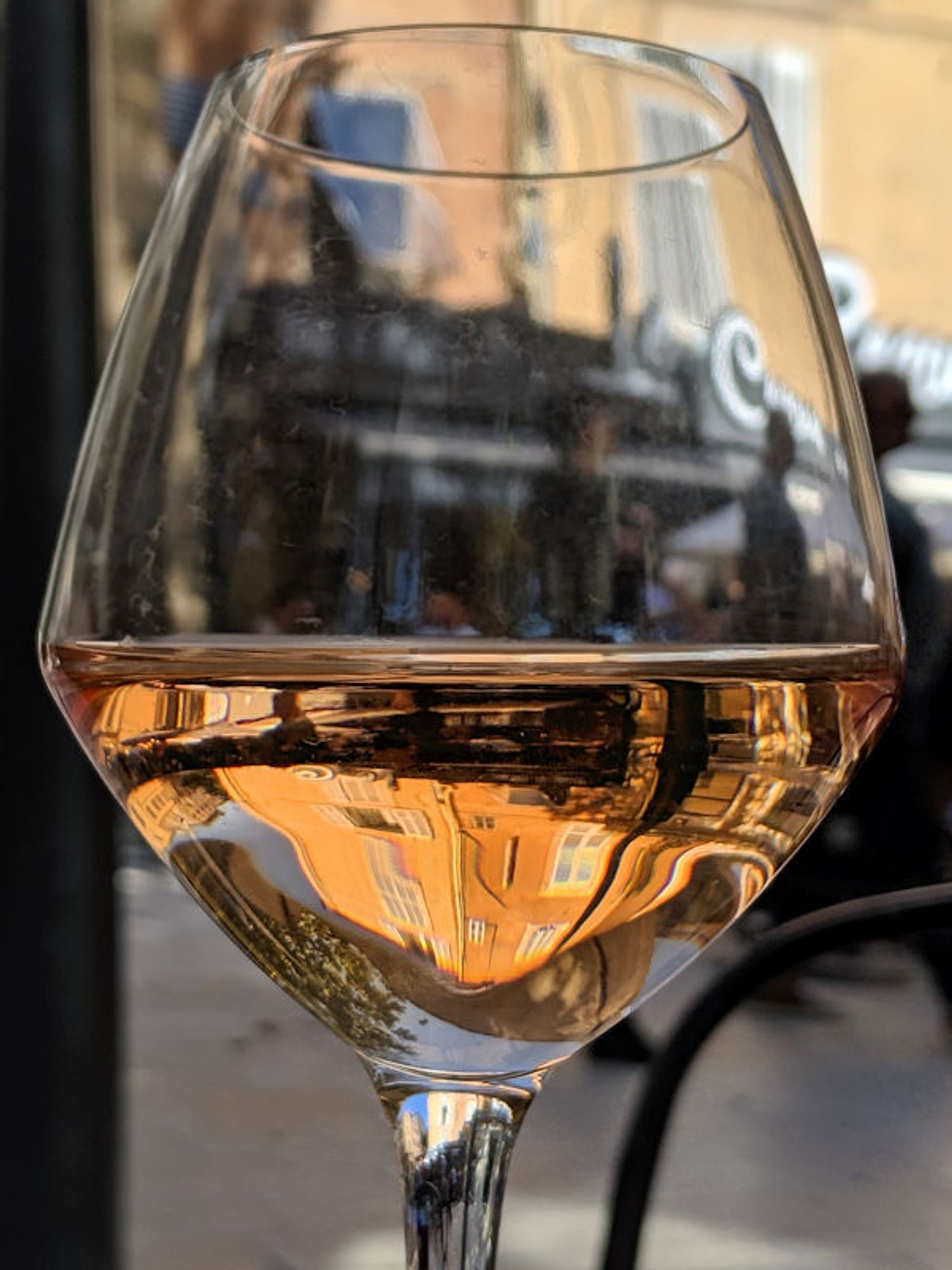 On the table at an outdoor café, the wine in half a wine glass reflects the buildings on one side of the plaza while the top part of the glass lets you see the buildings on the other side.