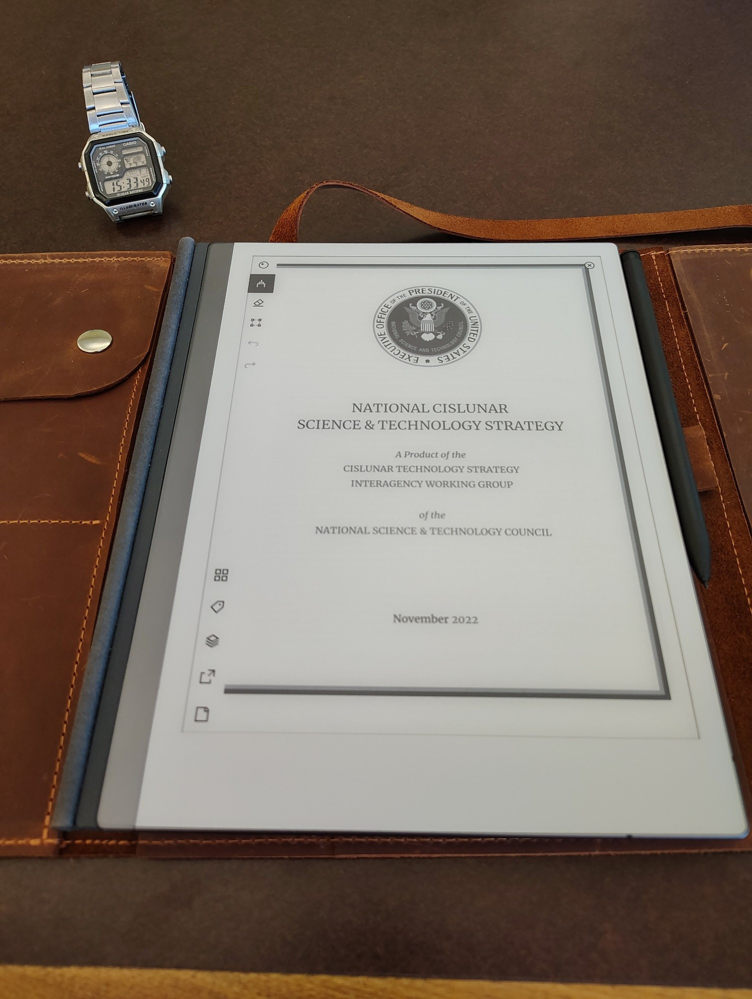 An e-ink reader open to the cover page of the NATIONAL SCIENCE & TECHNOLOGY STRATEGY report from November 2022.  Beneath it, a leather case for a notebook lays open.  Next to it, a Casio watch displays the time and thriftiness of its owner.