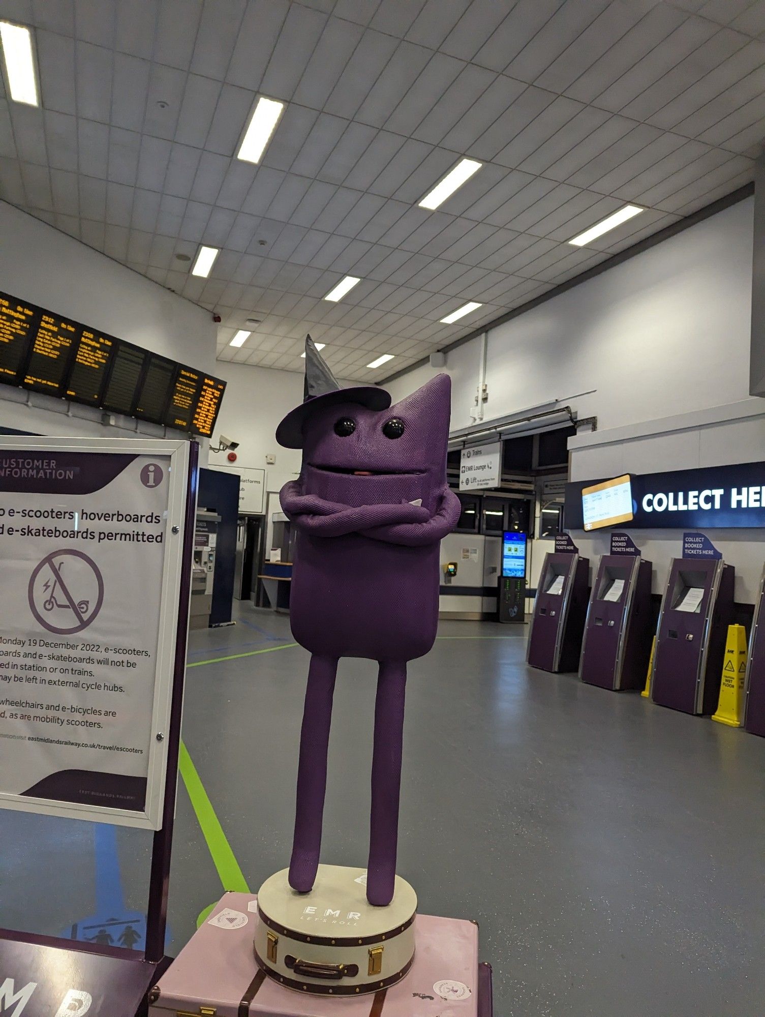 The EMR rail mascot in a witch/wizard hat standing in an empty train station with the ticket barriers in the background.