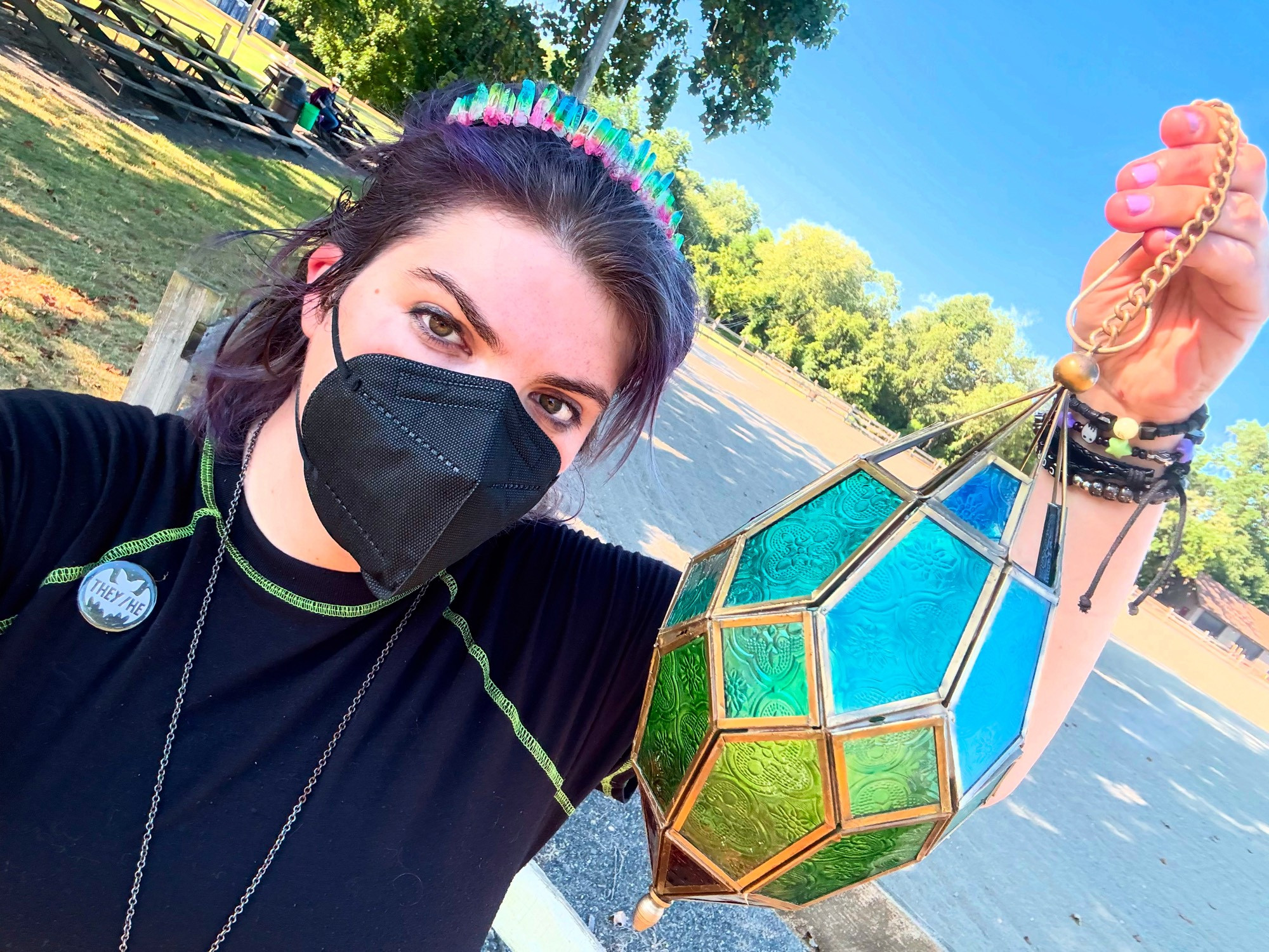 A light skinned person wearing a black dress, a tiara made of rainbow crystals, and a black KN95. They’re wearing a button that features the white outline of a bat and text that reads “They/ He”. They are holding a pendant shaped lantern made of green and blue glass. 