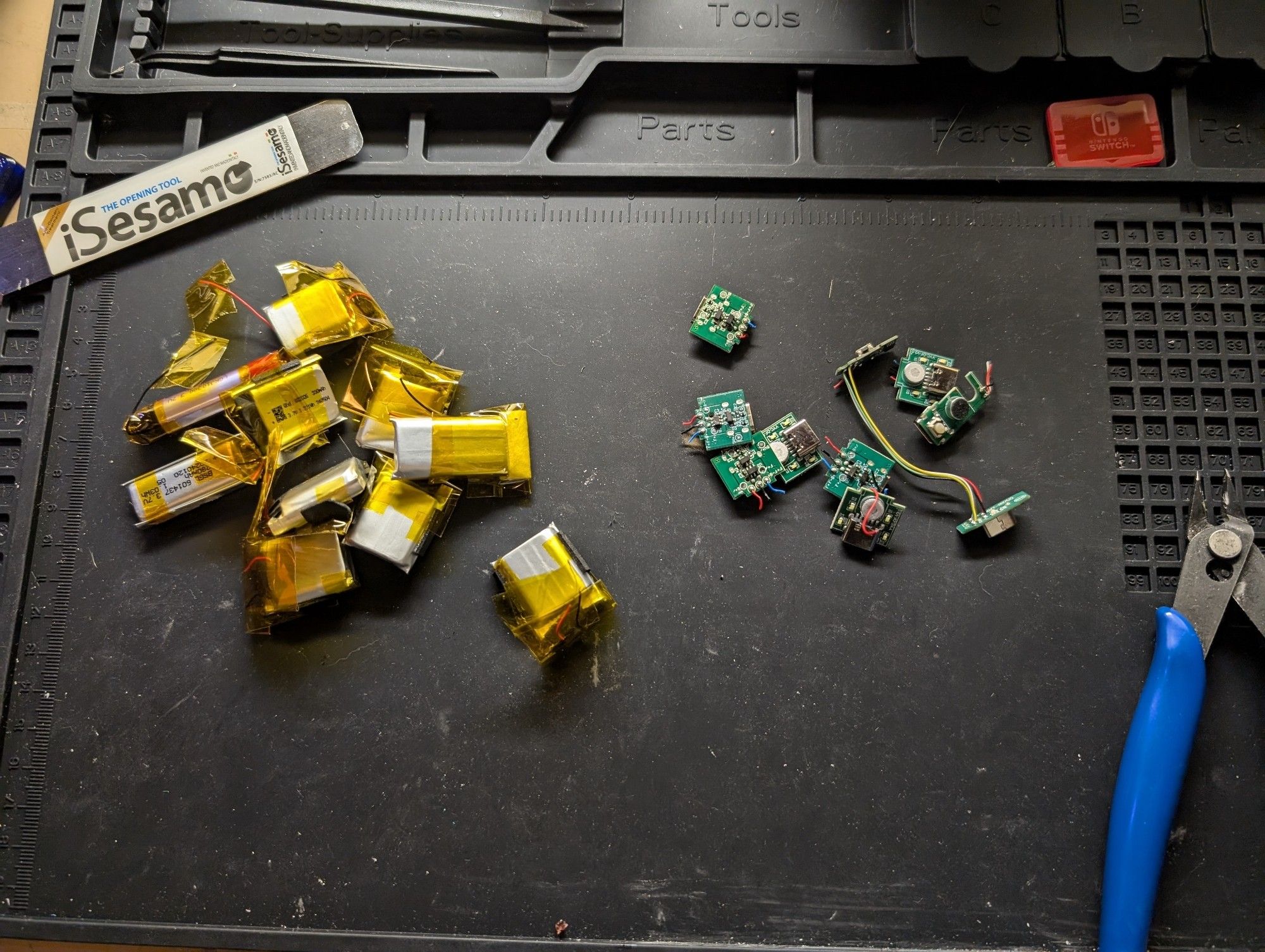 Photo of a workbench with a pile of small lithium rechargeable battery cells to the left of frame and a pile of their respective charging circuits.