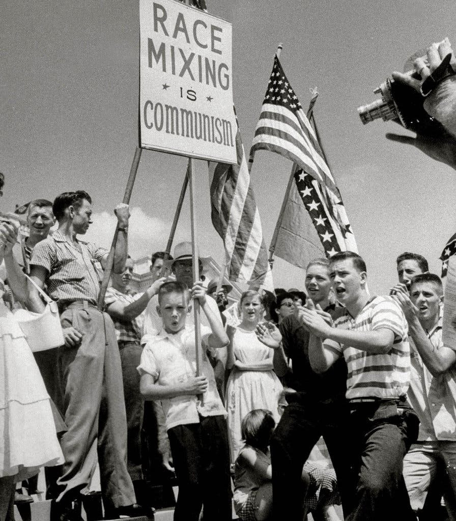 Angry white people protesting integration in Little Rock, Arkansas 1959.
One kid with a flattop haircut holds a  “race mixing is communism” sign. There’s an American flag, an Arkansas state flag and teenagers clapping and yelling.

A protest against school integration in Little Rock, Ark., in 1959.Credit...Francis Miller/Time & Life Pictures — Getty Images
