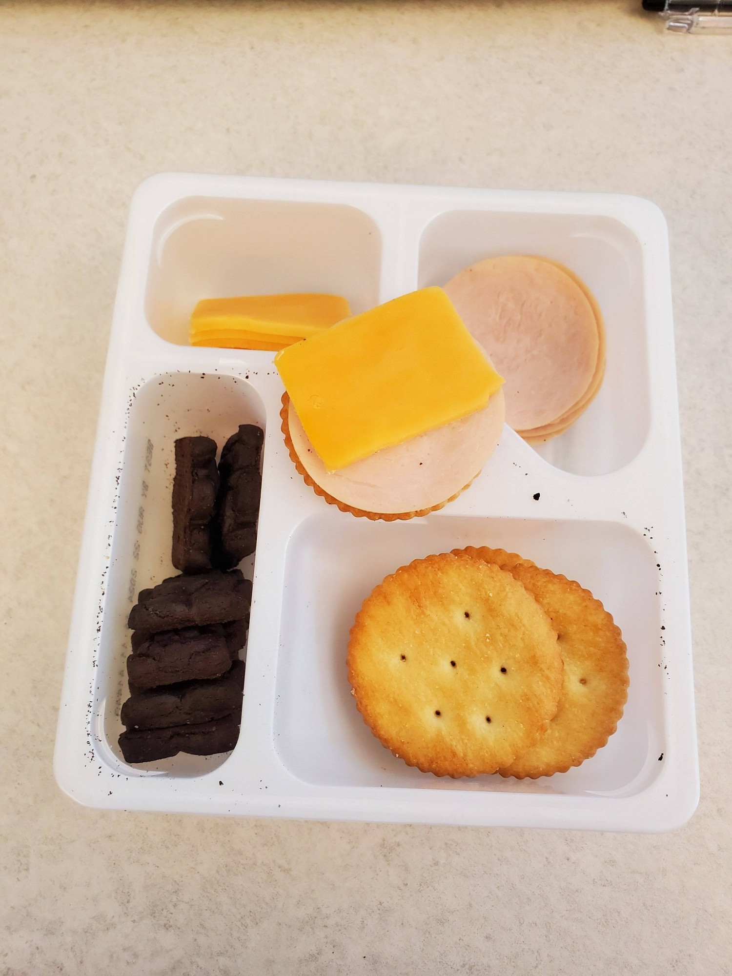 A small plastic tray with compartments. Clockwise from upper left: yellow cheese slices, little round turkey slices, generic buttery crackers and chocolate teddy bear cookies. On top of the tray is a cracker with turkey and cheese on it.