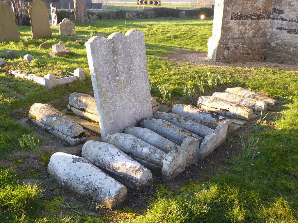 Centre: thirteen children’s graves with rounded casket shaped stones to top them in two rows, in front and behind their parent’s headstone in a grassy churchyard. A corner of the church is visible top right. Top left corner are other graves.