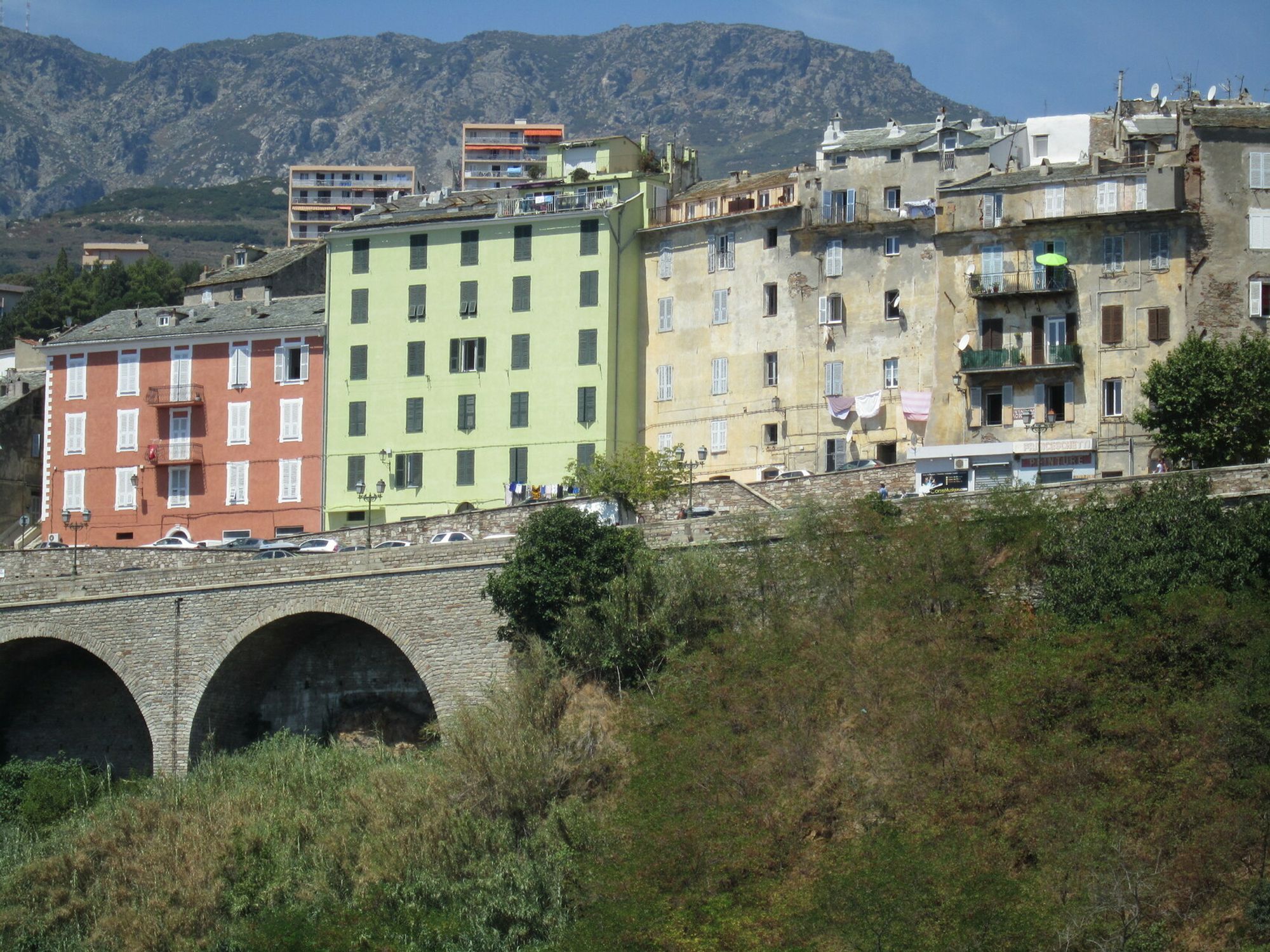 Maisons pittoresques en Corse, aux façades rouge, verte, grise, sur un pont moderne à arches, photo prise en 2013.