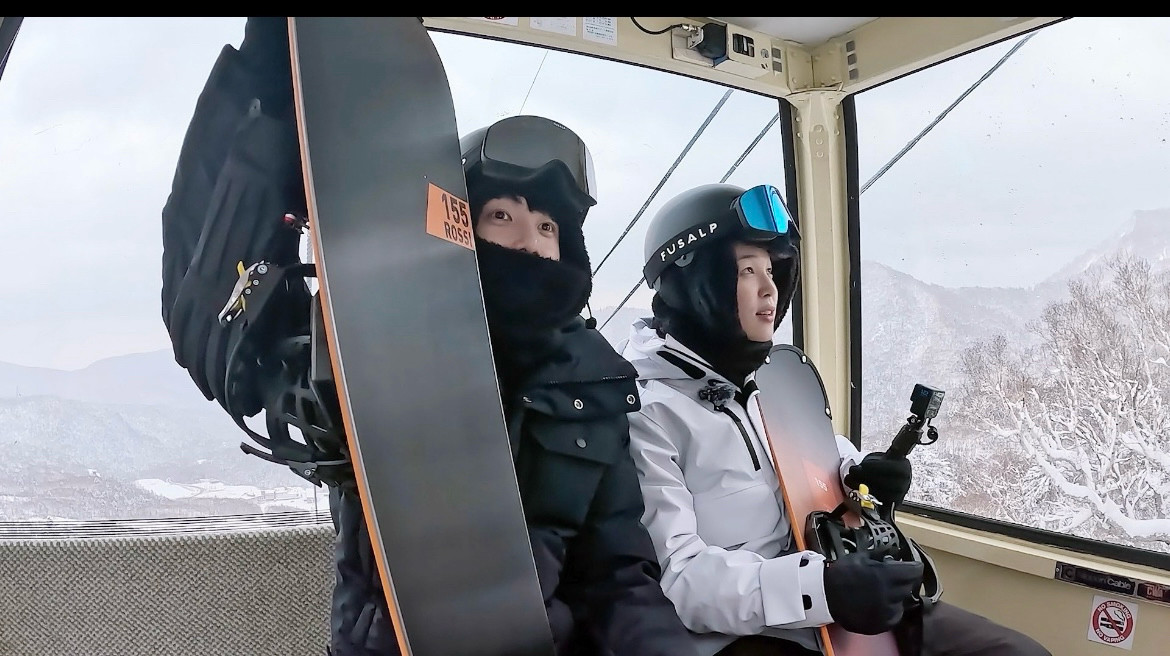 Jimin and Jungkook on a ski lift in snowboarding gear.