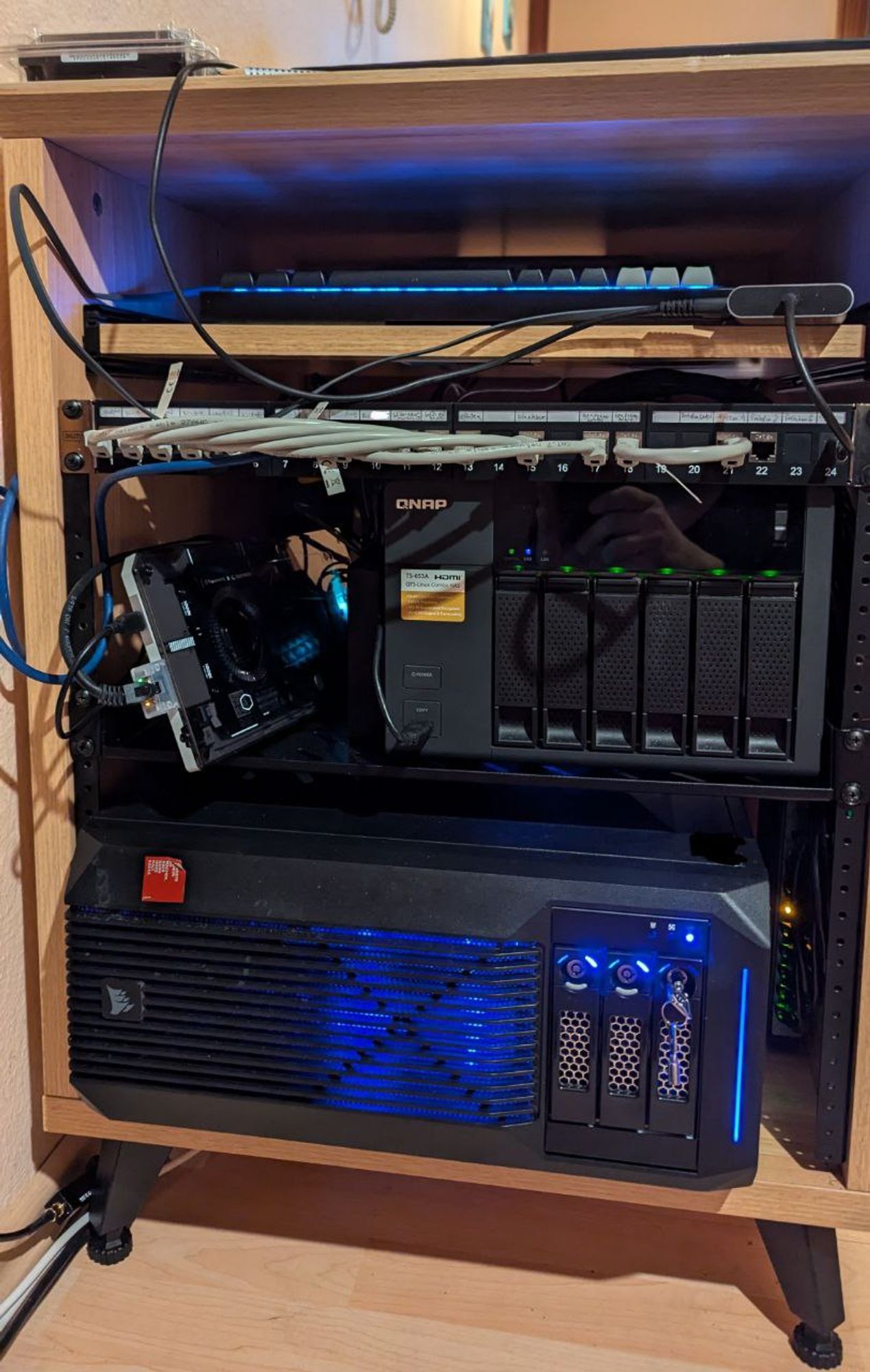 Front view of the wooden server rack with a cold spare drive on top of the rack, a blue-lit keyboard at the top-most shelf which is movable, a modular panel with lots of short CAT.7 cables and one USB-C cable at the right plugged into it, a Framework mainboard encased in a Cooler Master enclosure and a QNAP NAS in the middle, and a blue-lit PC in a Corsair SPEC-02 case with three of its 3.5" drive slots visible where two 5.25" slots would normally be.