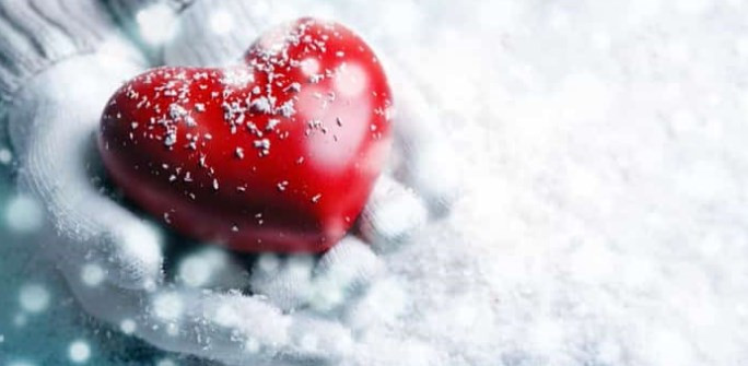 A shiny red heart laying in snow with a sprinkle of flakes on it.