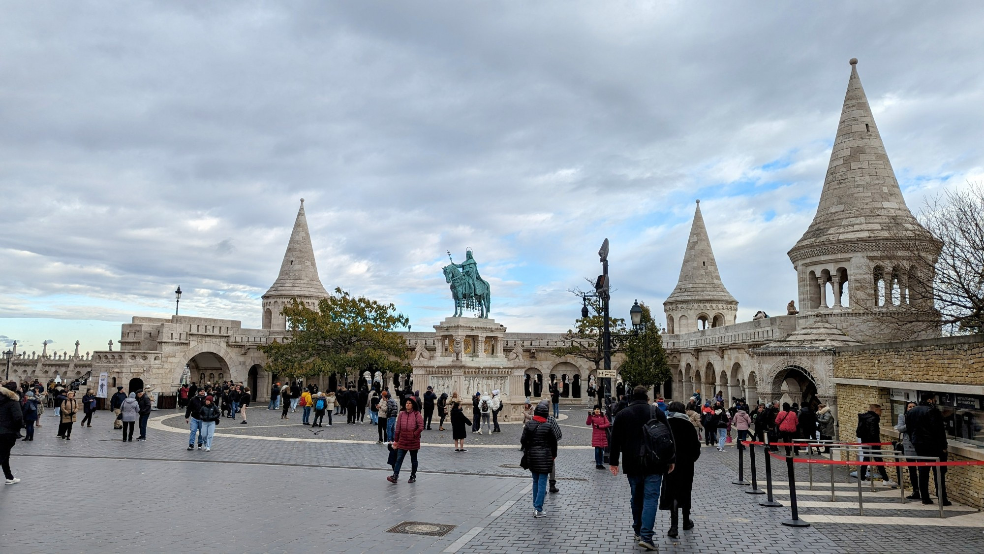 The photo shows the Fisherman's Bastion in Budapest, Hungary. It is a neo-Gothic style lookout terrace on the Buda side of the Danube river, offering panoramic views of the city. The photo captures the intricate architecture of the bastion, with its towers and statues, as well as the crowds of tourists exploring the site.
