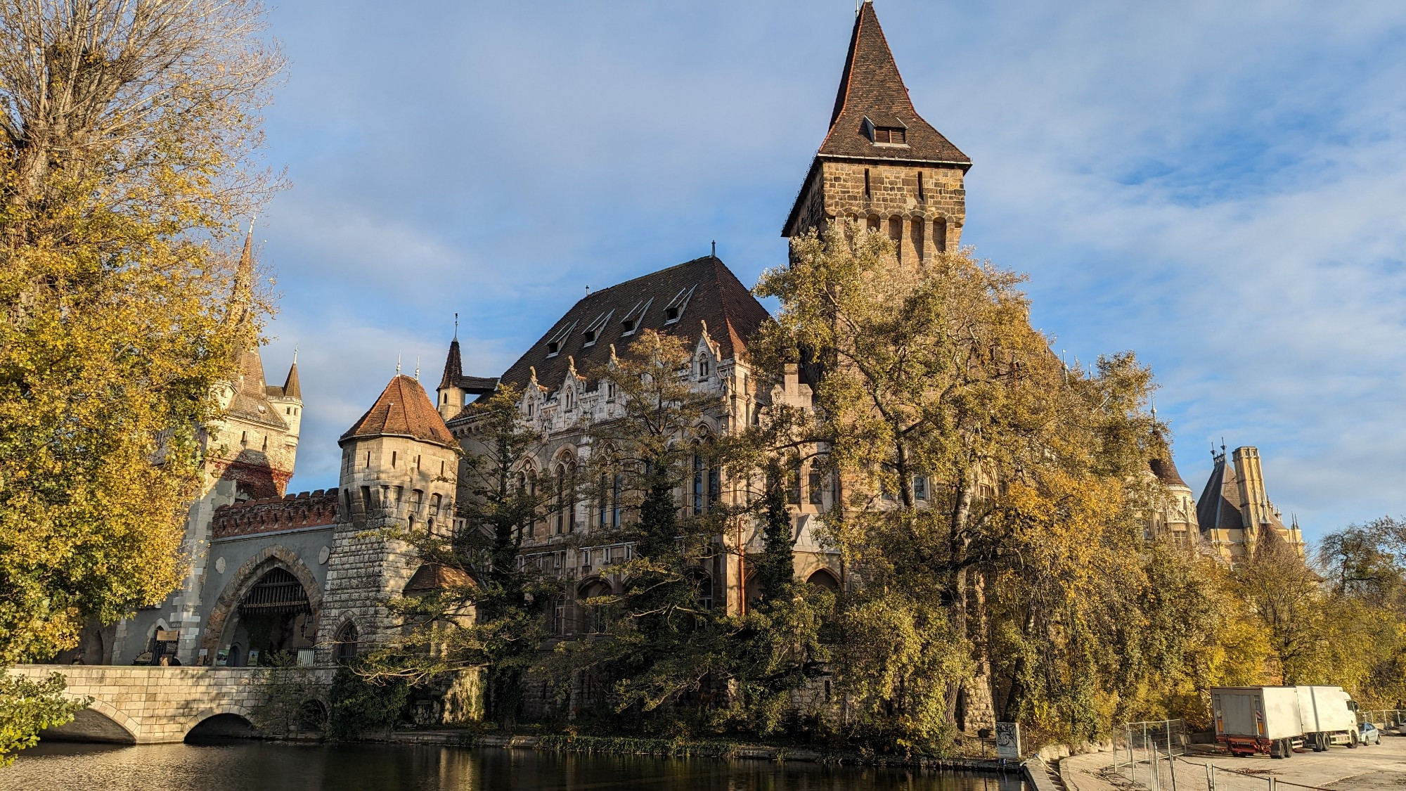 The photo shows Vajdahunyad Castle, a beautiful medieval-style castle in Budapest, Hungary. The castle is surrounded by a moat and is reflected in the water. The sky is blue and the trees are green.
