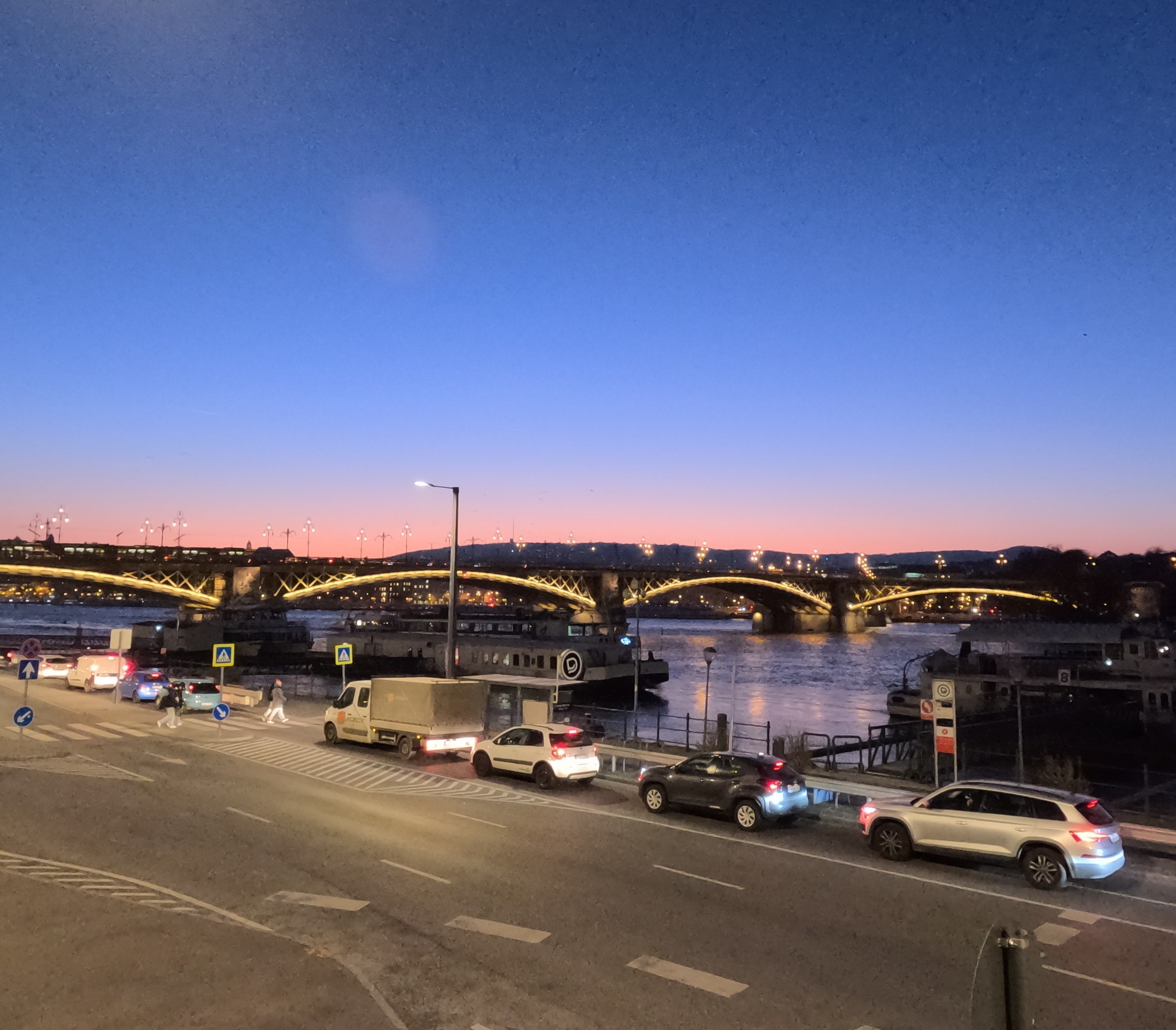 The photo shows a city street at night. There are cars parked on both sides of the street, and a Danube river runs along one side. The sky is dark, with a few lights visible in the distance. A bridge can be seen crossing the water.
