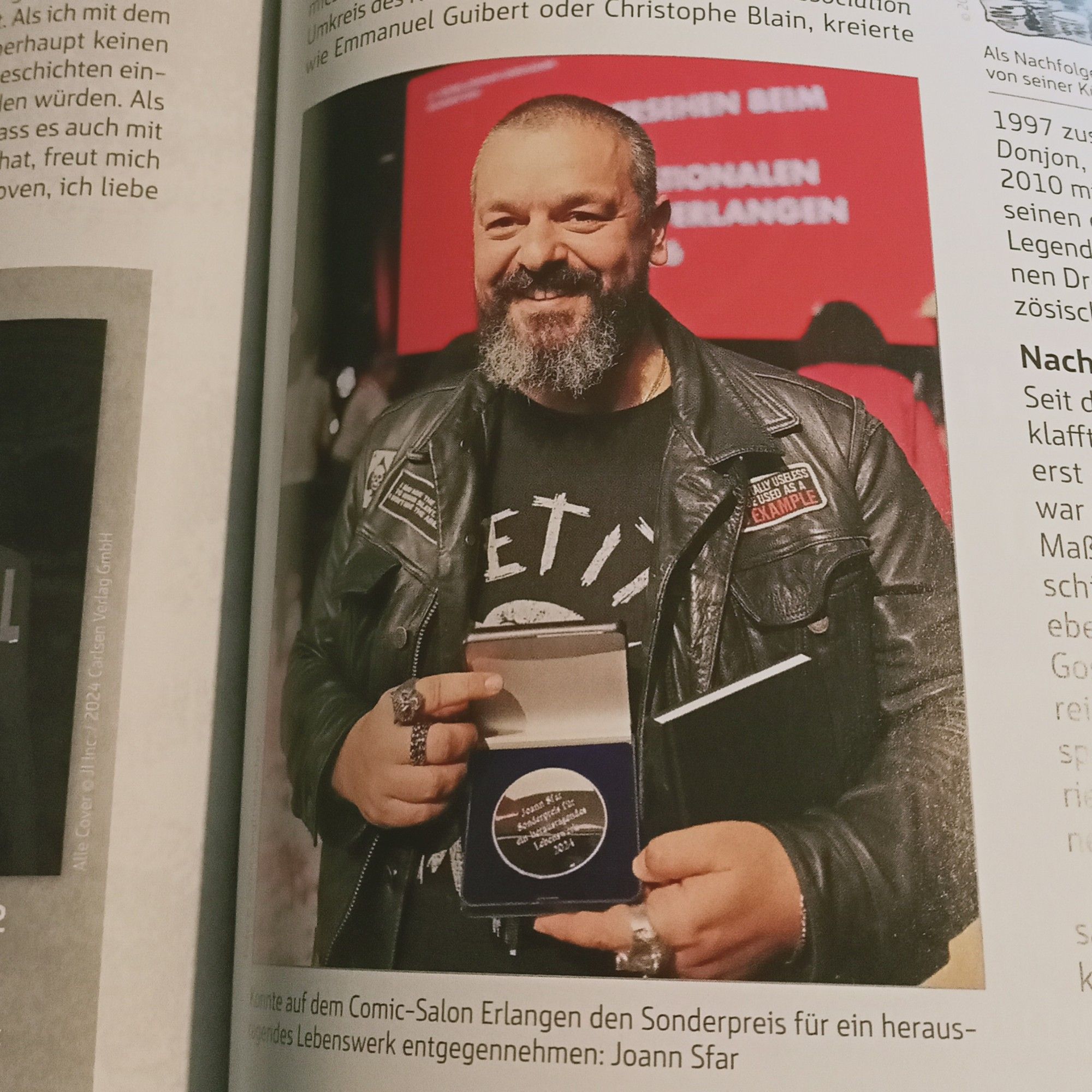 Knuffiger untersetzter Mann mit längerem graumelierten Vollbart zeigt stolz lächelnd seinen Preis für das Lebenswerk. Er trägt eine schwarze Lederjacke mit Aufnähern über einem schwarzen Motiv-Shirt, und an seinen Fingern stecken mindestens drei fette silbrige Ringe.